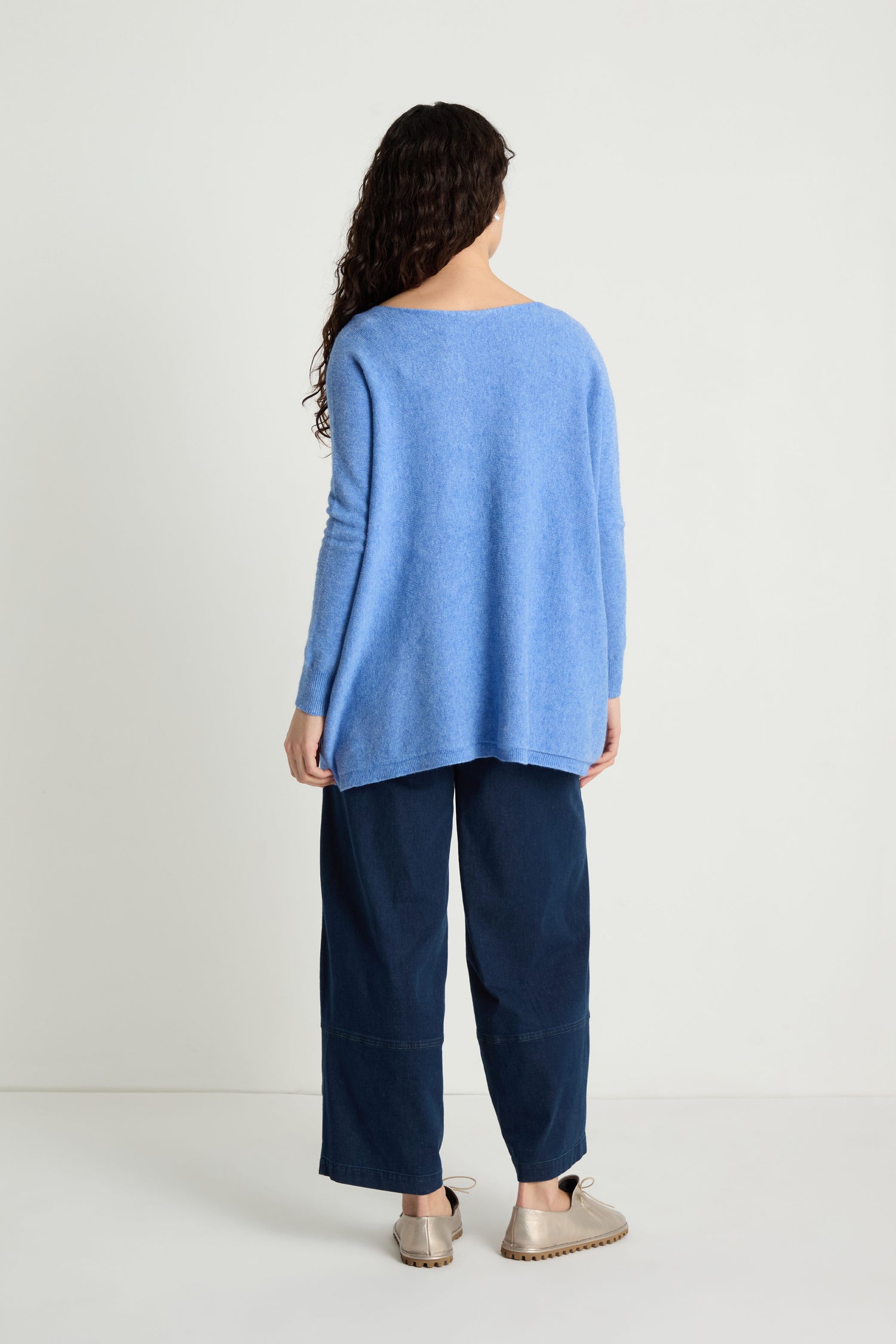 A person with long curly hair faces away, wearing the Two Pocket Oversize Knit in blue from women's knitwear, paired with wide dark blue pants and beige sandals against a plain white background.