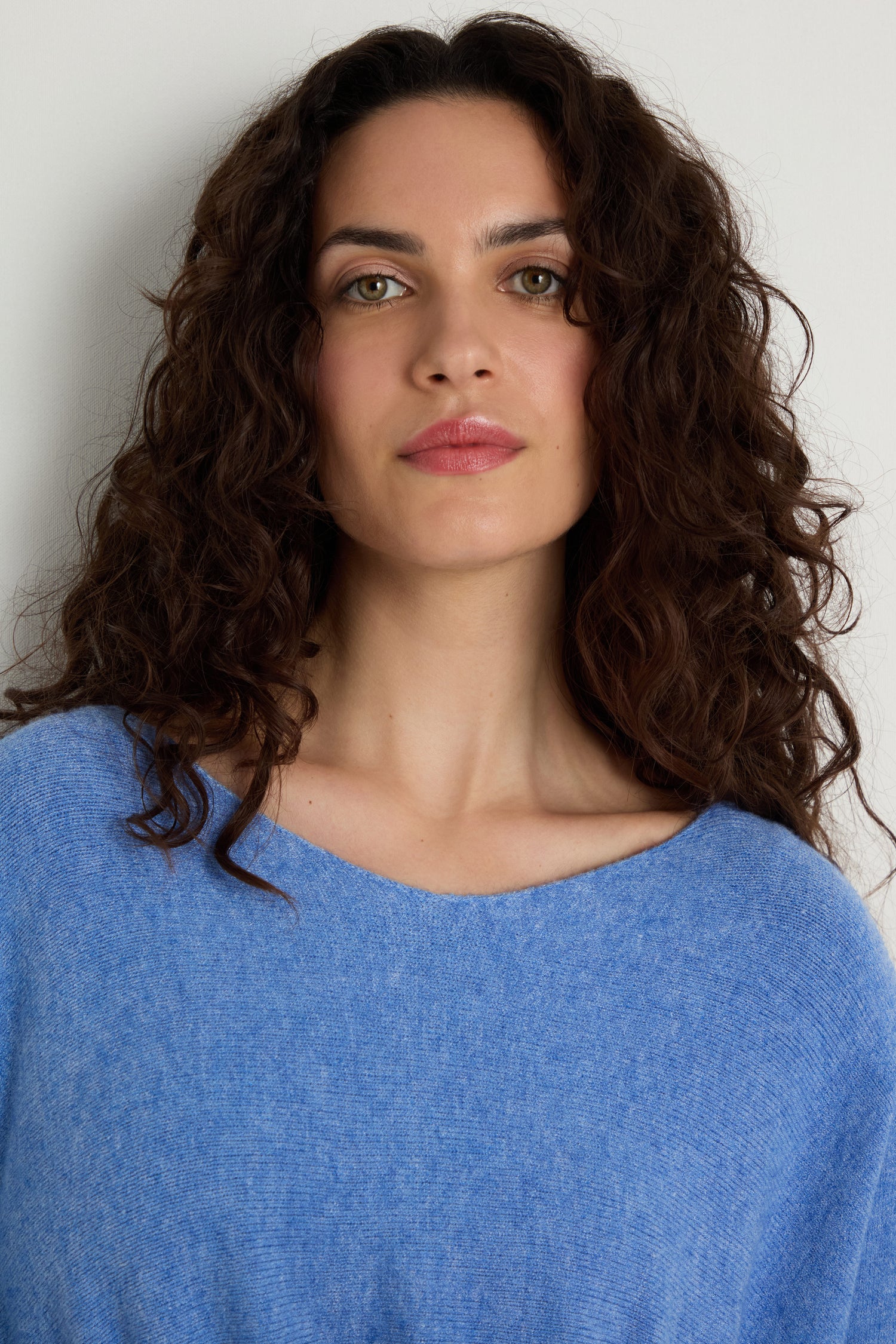A woman with long curly brown hair wears the Two Pocket Oversize Knit in light blue, standing against a plain white background and looking directly at the camera.