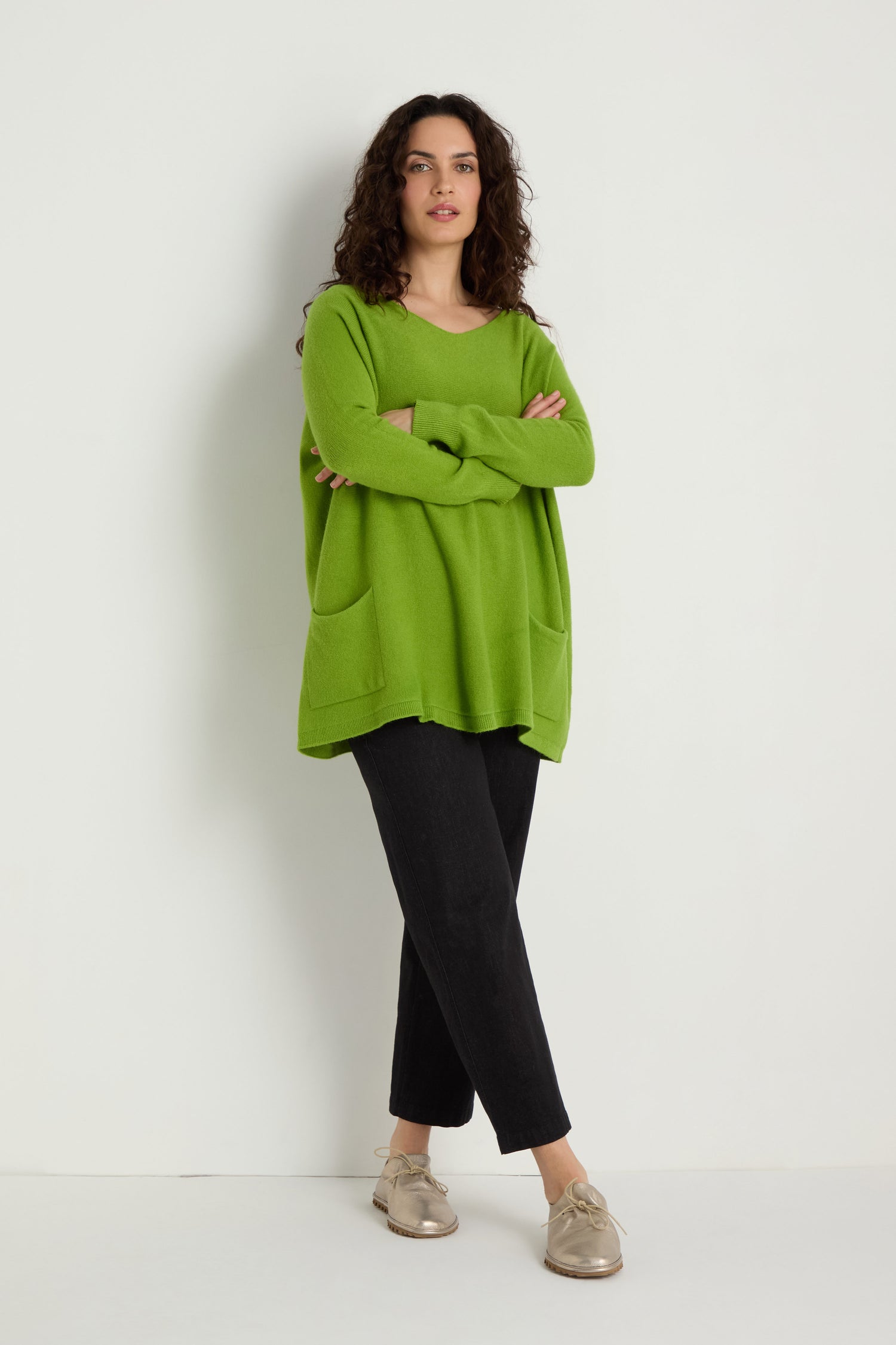 A person stands against a plain white background, wearing the Two Pocket Oversize Knit in green from women's knitwear, paired with black pants and beige shoes, with arms crossed over their chest.