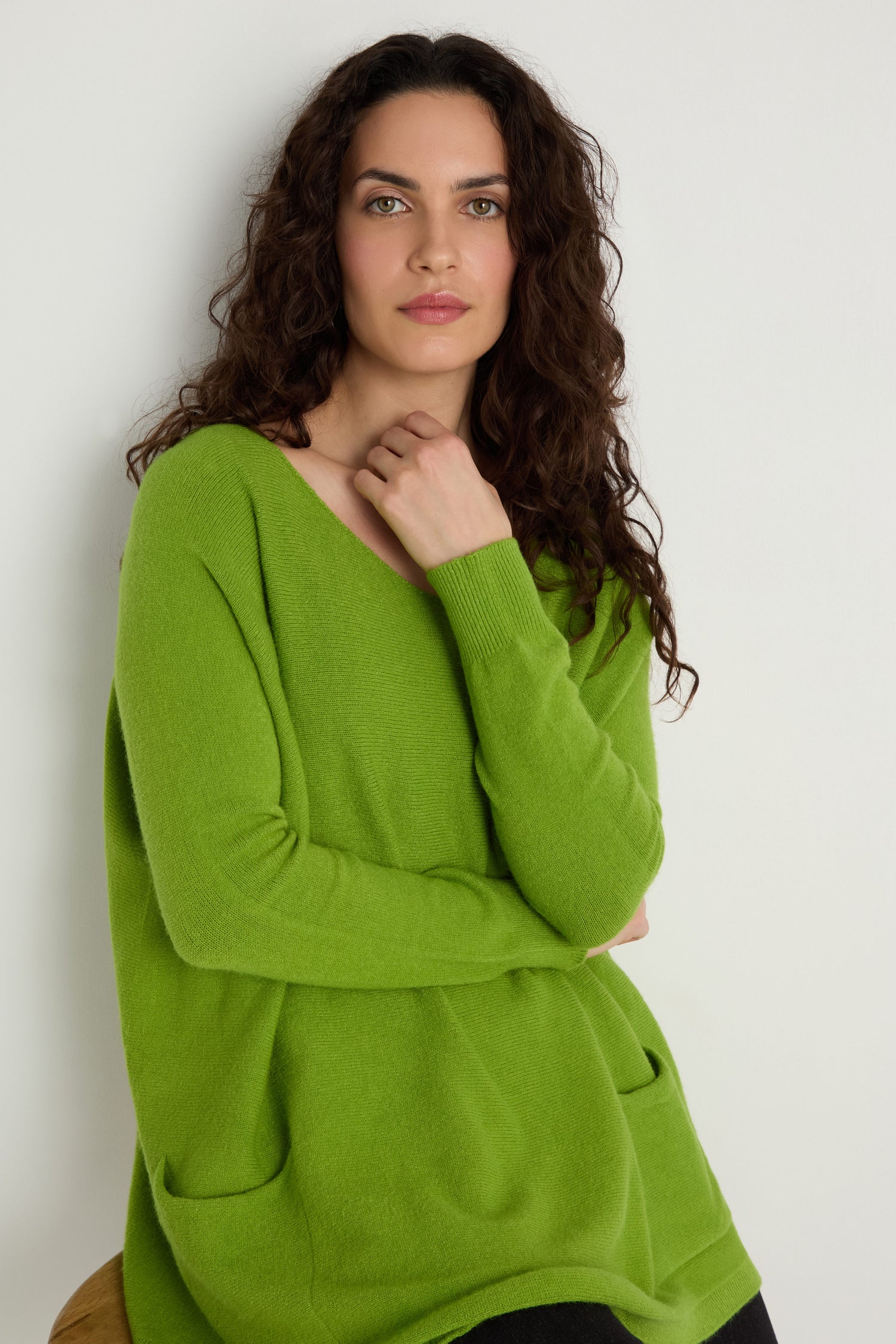 A woman with long curly brown hair models the Two Pocket Oversize Knit in green, standing against a white wall and looking upward with one hand on her chin—ideal for fans of women’s knitwear.
