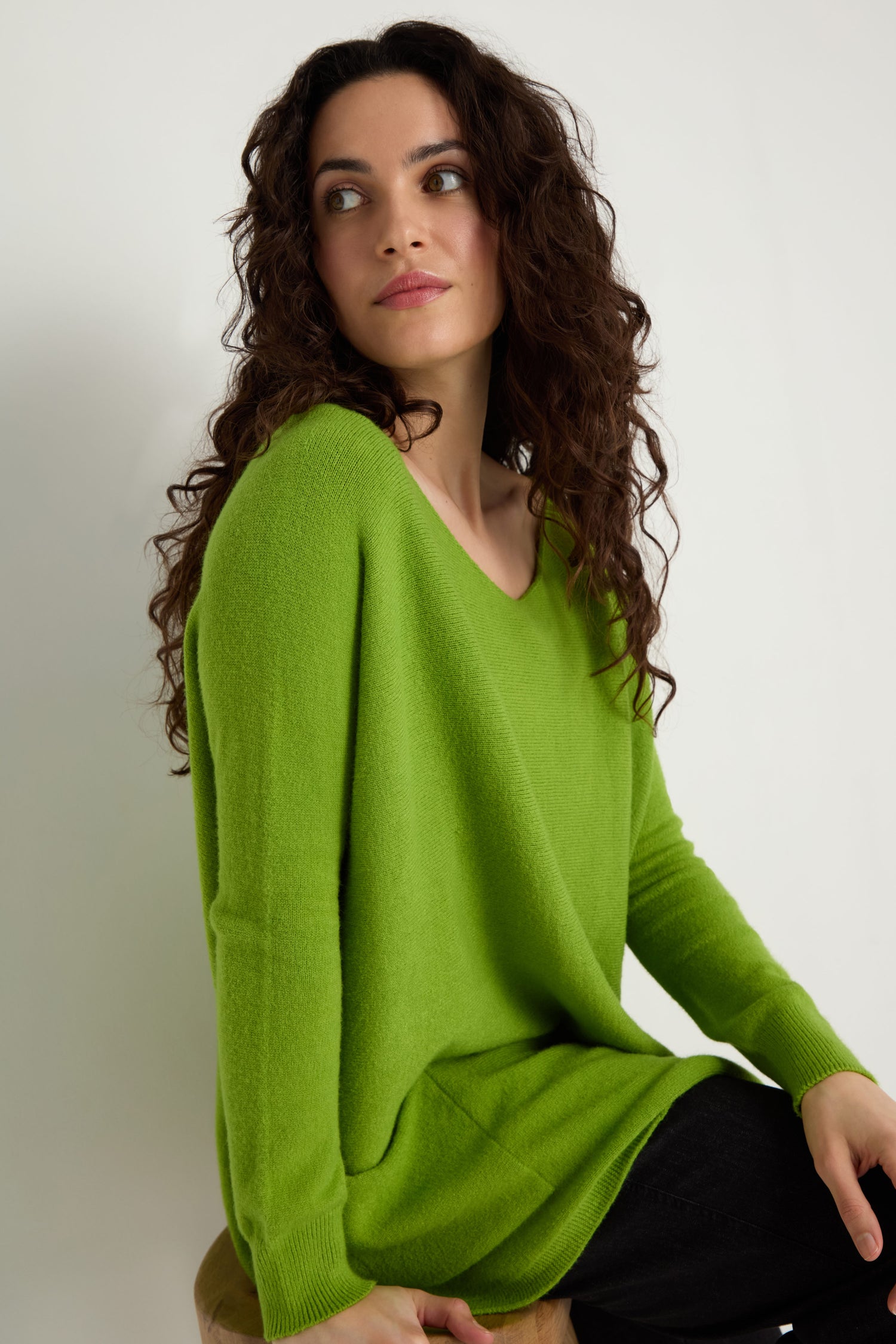 Woman with long curly hair wearing the Two Pocket Oversize Knit in bright green and black pants sits on a stool, looking to the side against a plain white background—showcasing chic women’s knitwear.