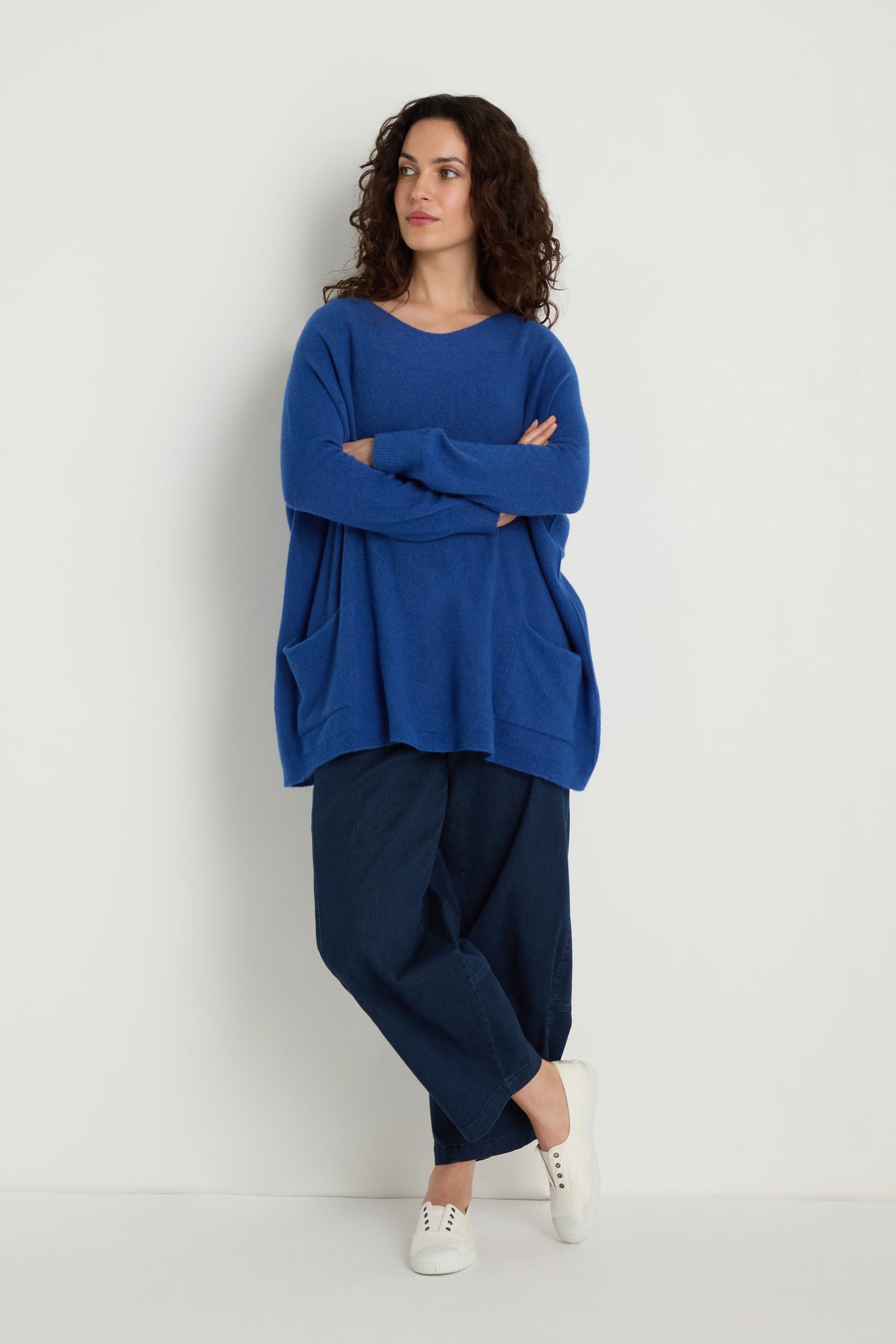 A person stands against a plain background, wearing the Two Pocket Oversize Knit in blue from women's knitwear, paired with dark pants and white sneakers. Their arms are crossed and they're looking to the side.