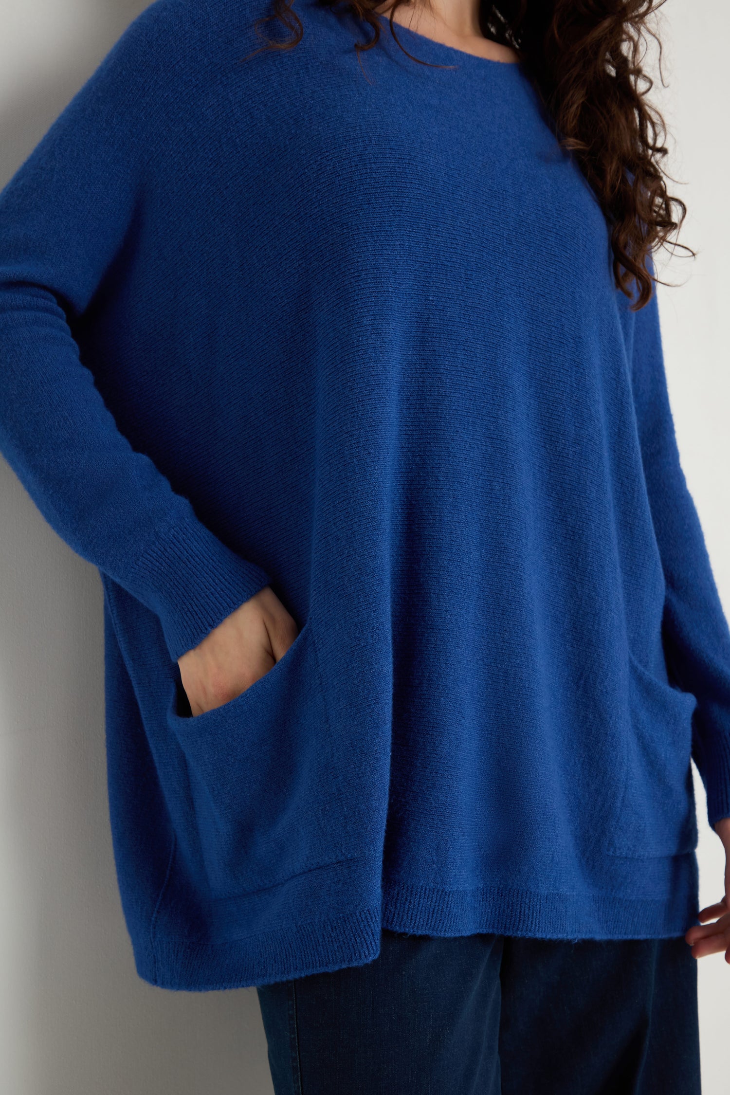A person wearing the Two Pocket Oversize Knit—an oversized blue women’s jumper with long sleeves and two front pockets—paired with dark pants, stands against a plain background.
