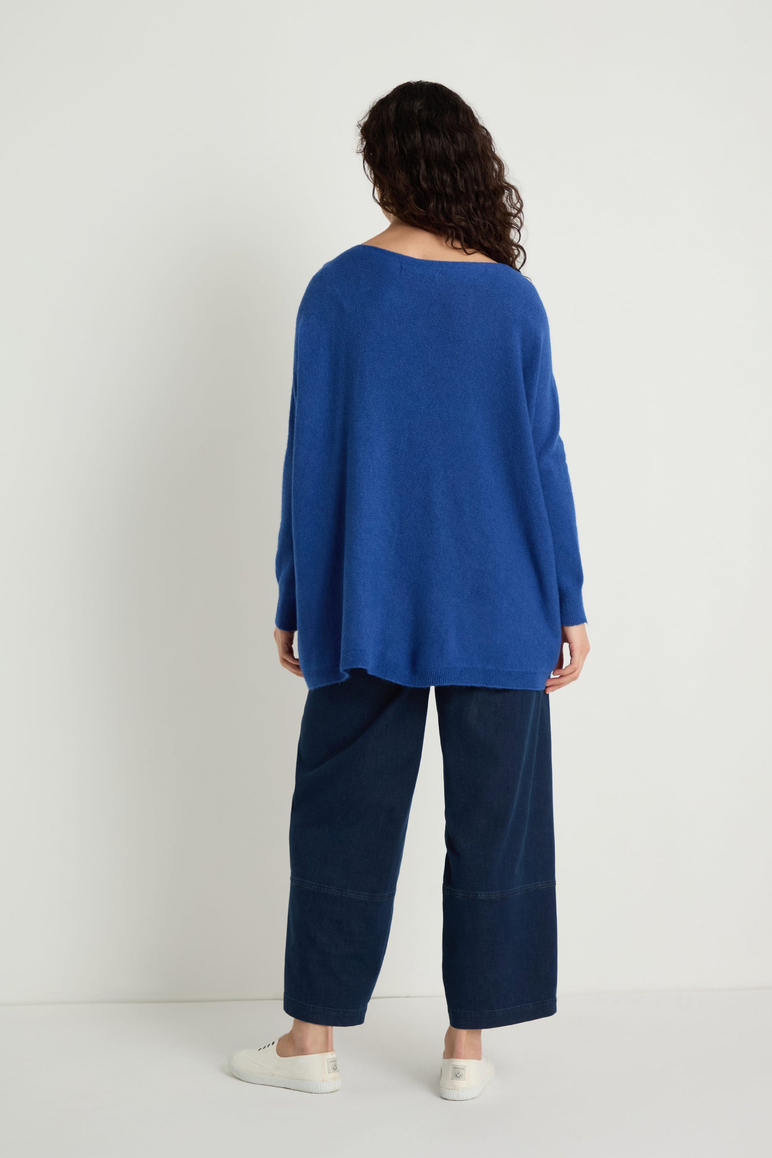 Person standing with their back to the camera, wearing the Two Pocket Oversize Knit in blue—classic women’s knitwear—paired with wide-leg dark blue pants and white sneakers against a plain white background.