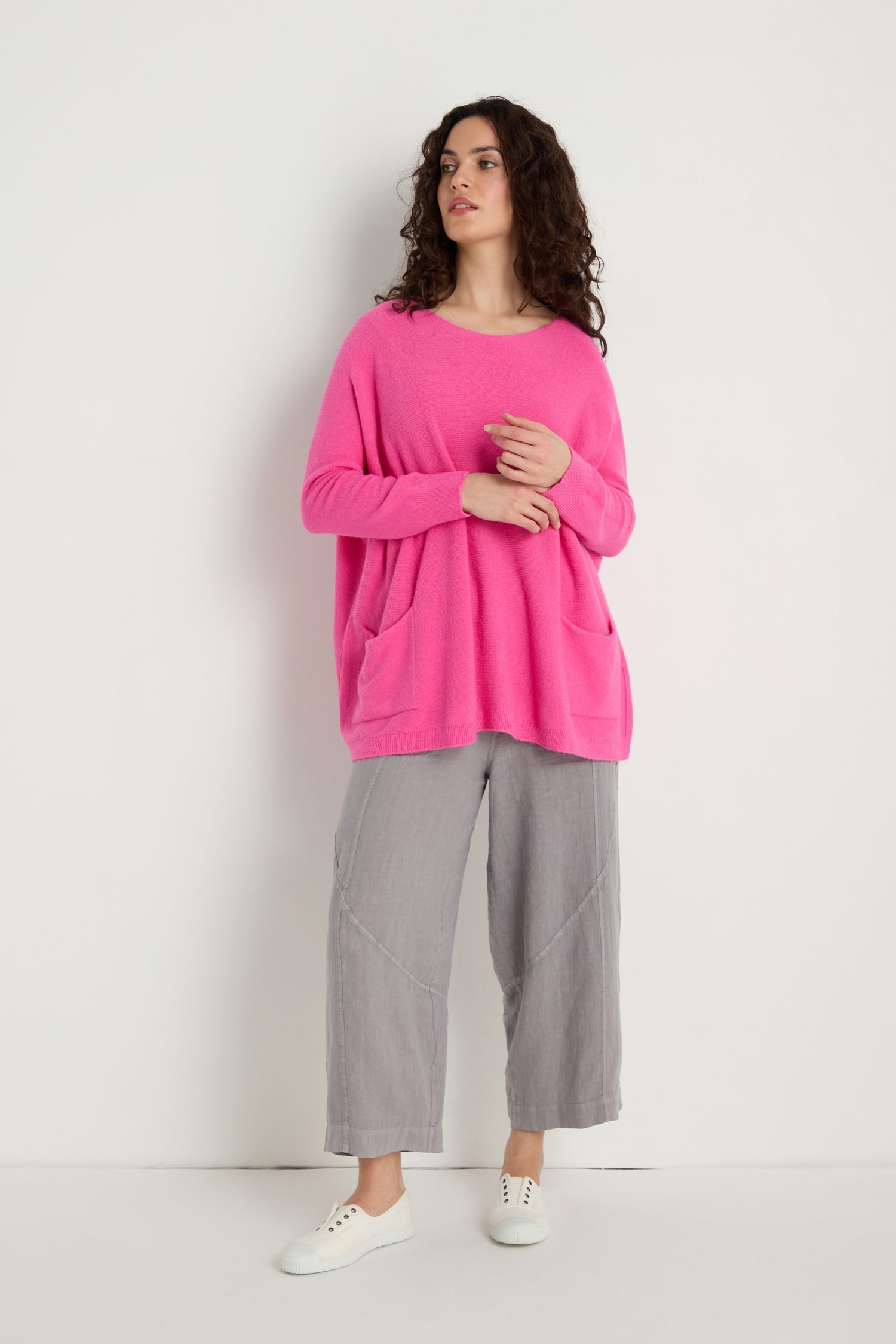 A woman stands against a white wall wearing the Two Pocket Oversize Knit in bright pink, paired with light gray wide-leg pants and white sneakers.
