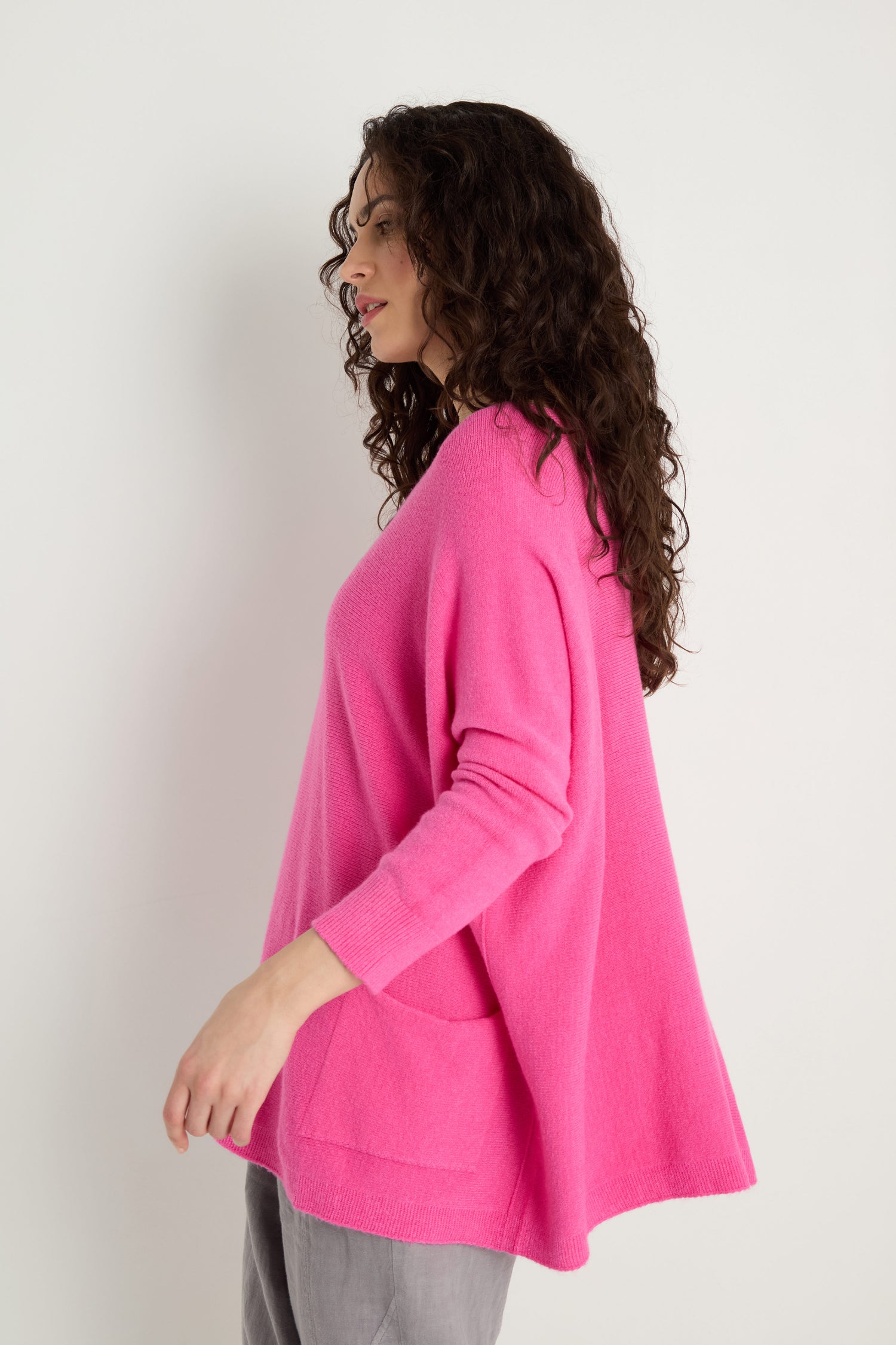 A woman with curly hair models the Two Pocket Oversize Knit in bright pink with gray pants, highlighting stylish women's knitwear against a plain white background.