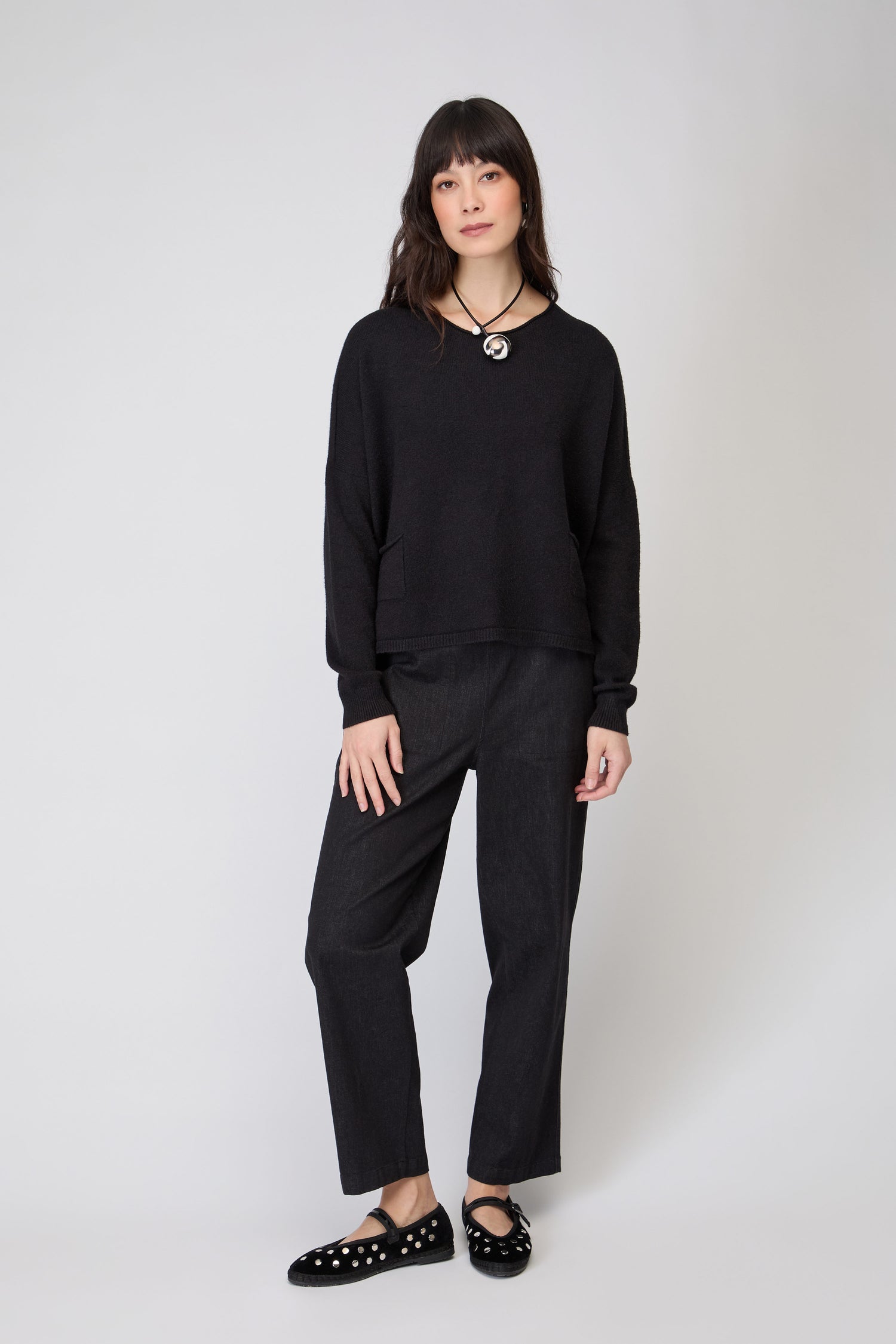A woman stands against a plain background, wearing the Two Pocket Black Knit sweater, black pants, black shoes with white dots, and a circular pendant necklace.