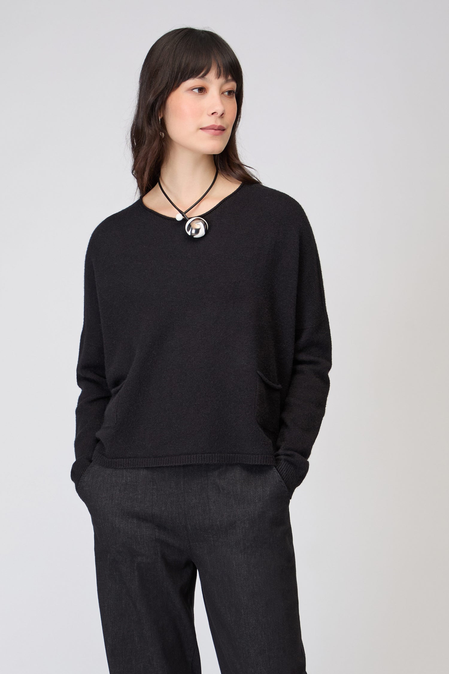 Wearing the Two Pocket Black Knit, a boxy black top, paired with dark pants and a round pendant necklace, the person stands with hands in pockets against a plain background.