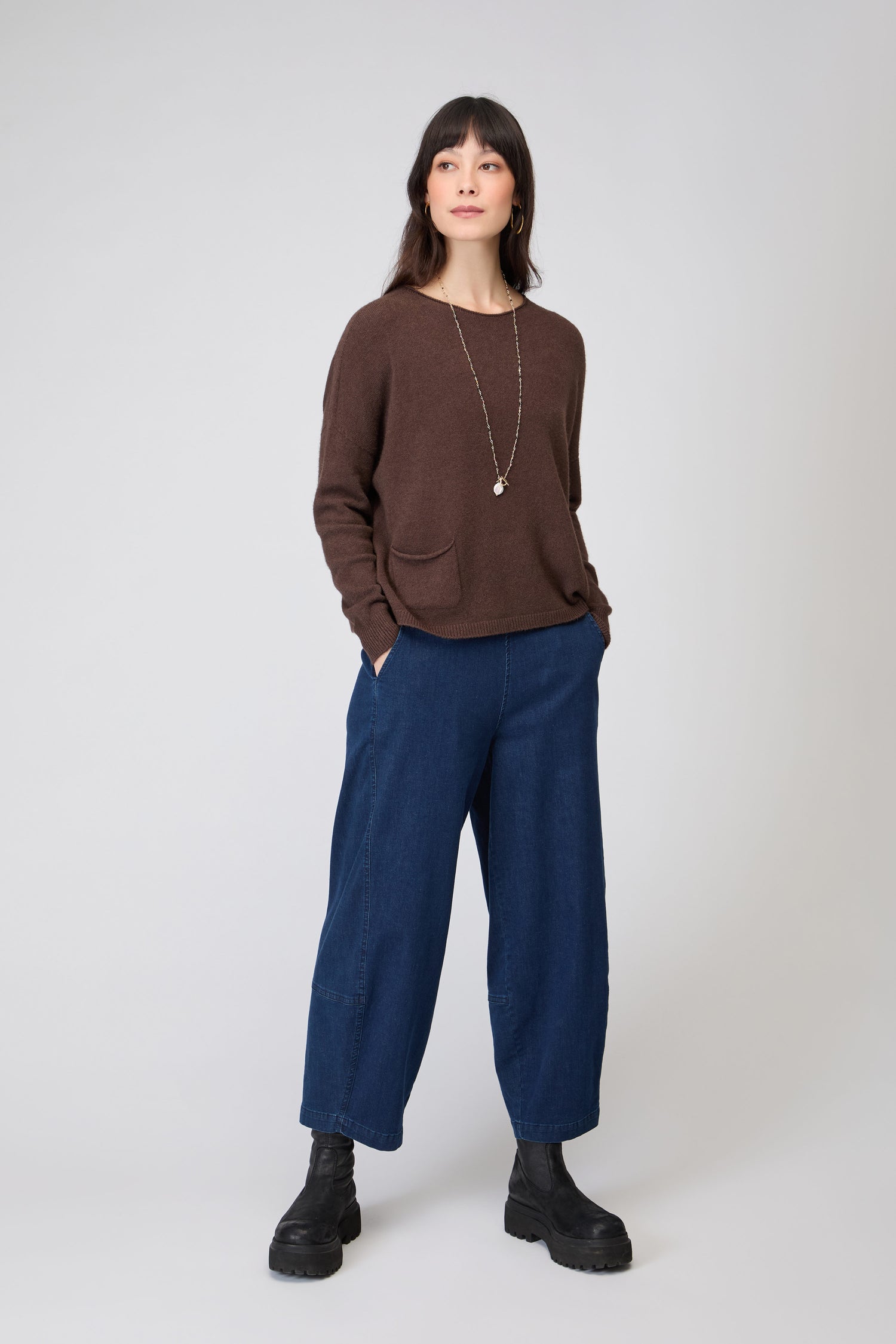 Woman standing in a relaxed pose, wearing the Two Pocket Knit brown sweater, wide-leg blue jeans, black boots, and a long necklace against a plain light background.