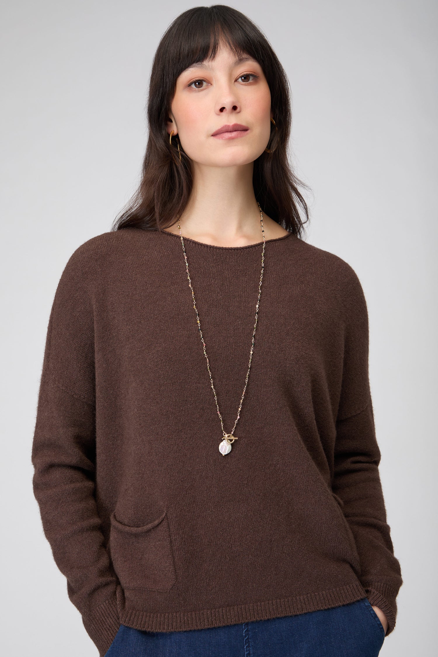 A woman with long dark hair wears the Two Pocket Knit, featuring a viscose-rich fabric and front pockets, paired with blue pants and a pendant necklace, standing against a plain light gray background.