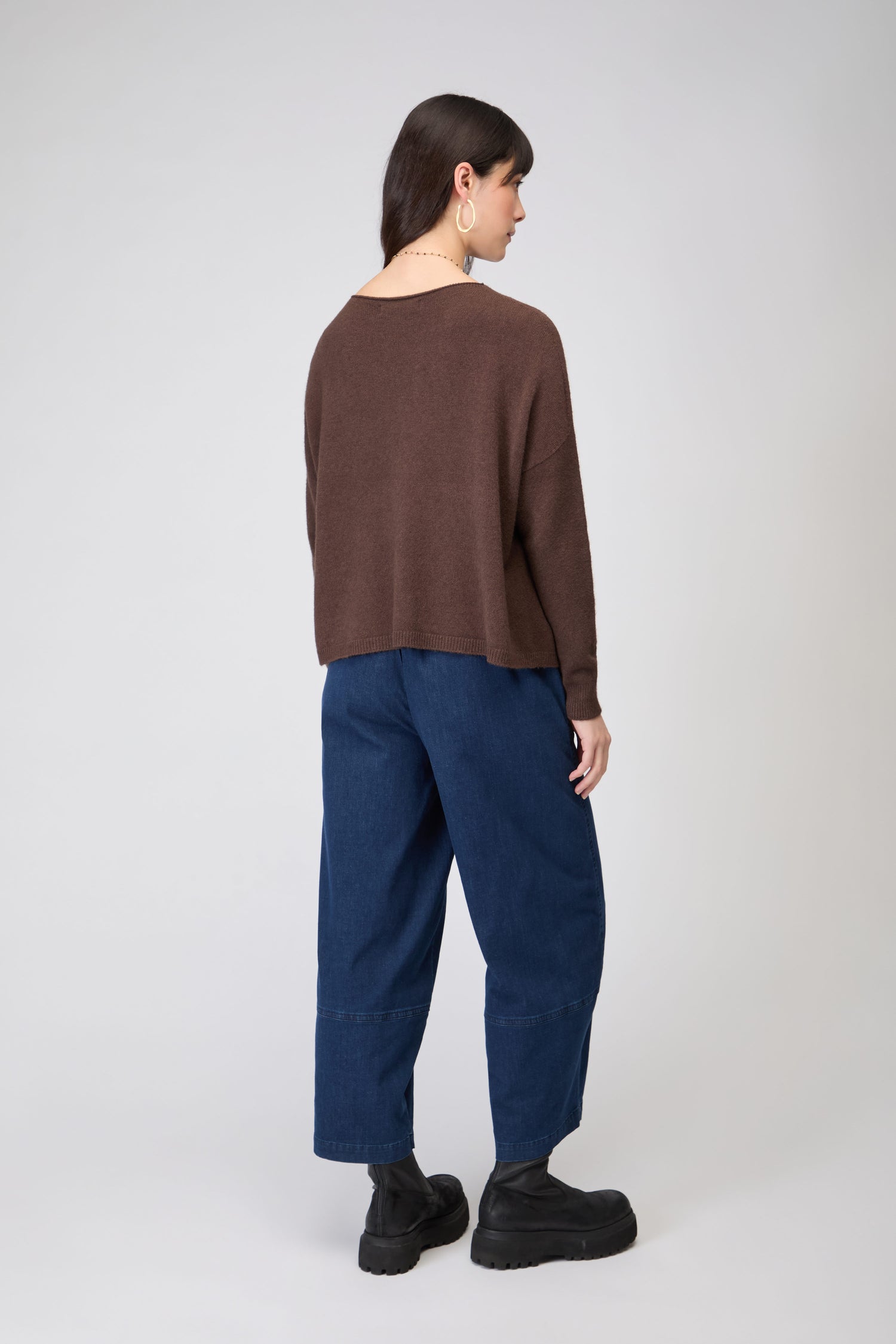 A woman faces away in a relaxed, timeless look featuring the Two Pocket Knit—a loose brown women’s jumper—paired with wide blue jeans, black boots, hoop earrings, and a necklace against a plain light background.