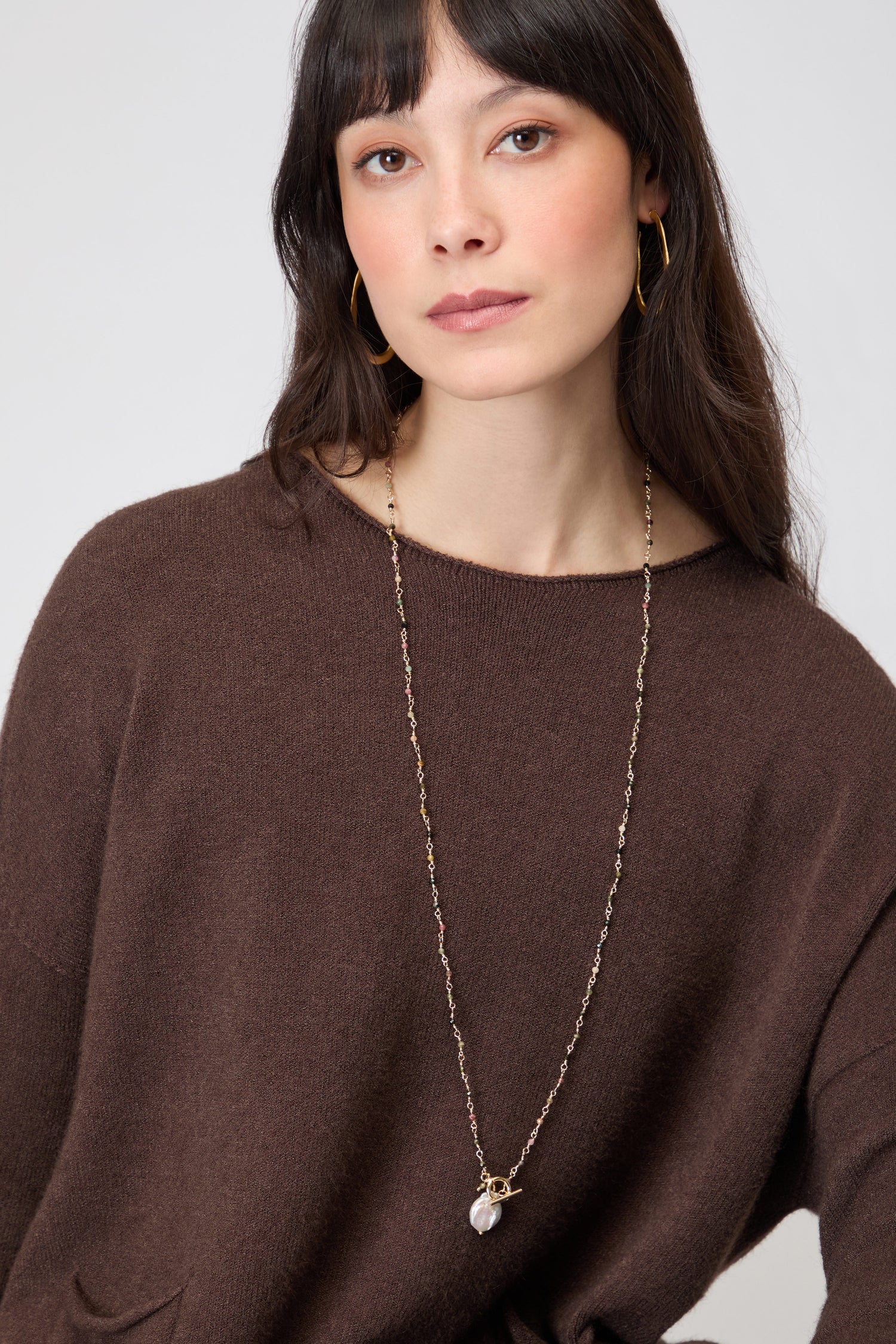 A woman with long brown hair wears the Two Pocket Knit jumper, gold hoop earrings, and a long gold necklace with a pearl pendant. She looks slightly to the side.
