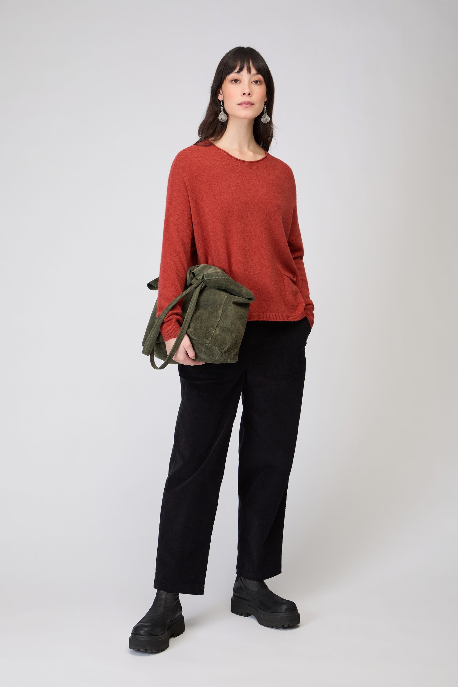 A woman stands against a plain background wearing a red Two Pocket Knit jumper, black pants, black boots, and carrying an olive green shoulder bag.