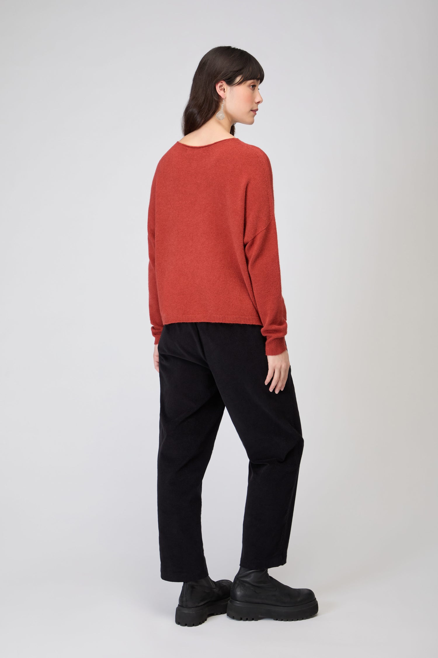 Woman standing on a white background, facing away, wearing a red Two Pocket Knit jumper, black pants, and black platform shoes.