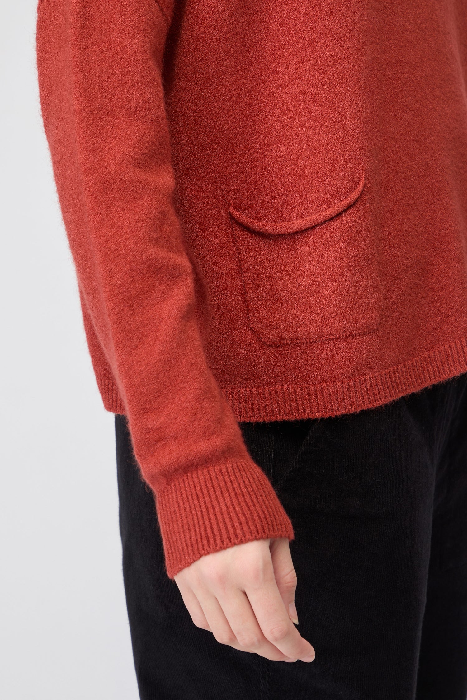 Someone in a red Two Pocket Knit women’s jumper with black pants, shown from shoulder to mid-thigh, arm relaxed at their side—perfect for lovers of timeless, comfortable knitwear.