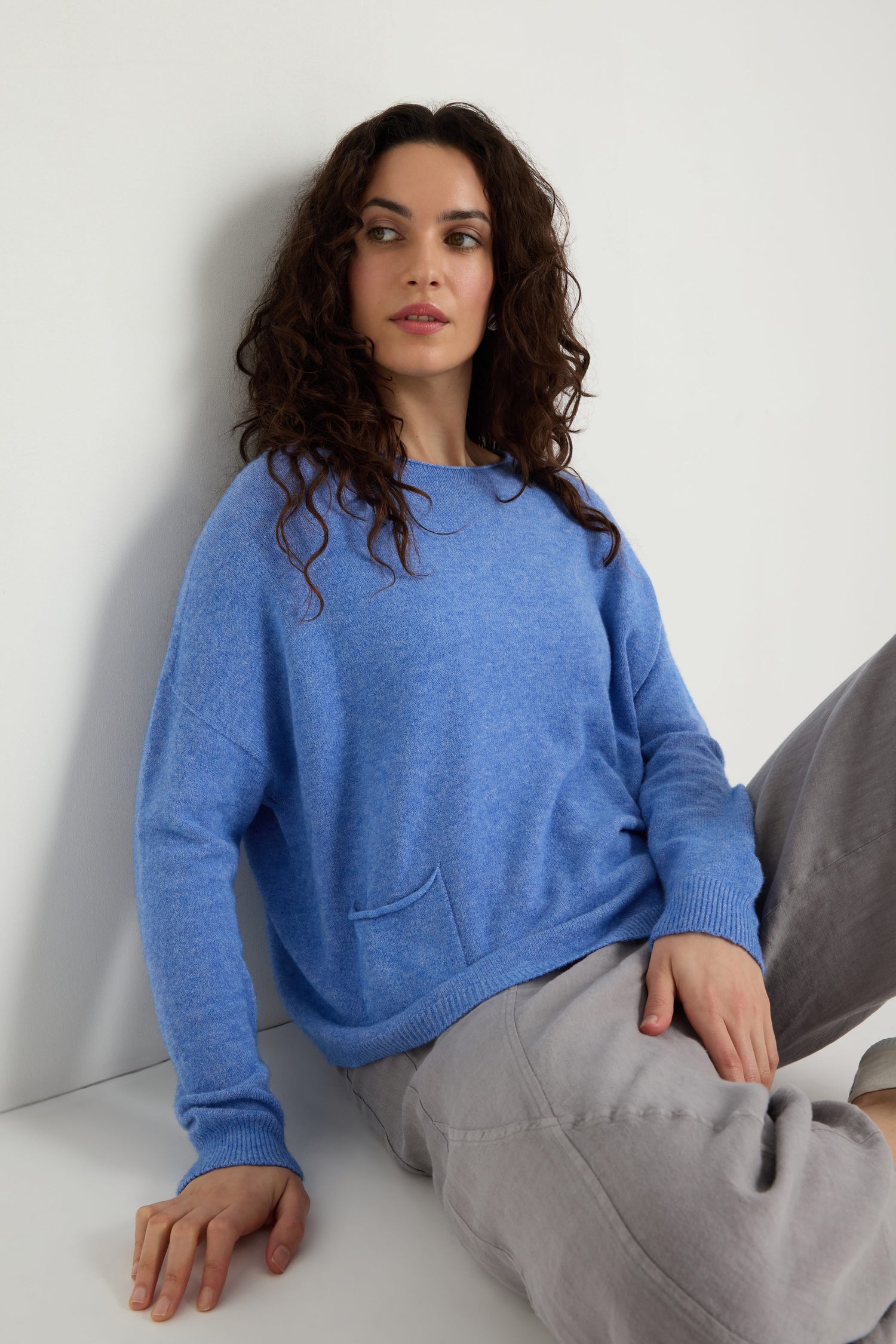 A woman with curly brown hair wears the Two Pocket Knit sweater in blue with gray pants, sitting against a white wall and looking slightly to the side—an ideal look for everyday wear.