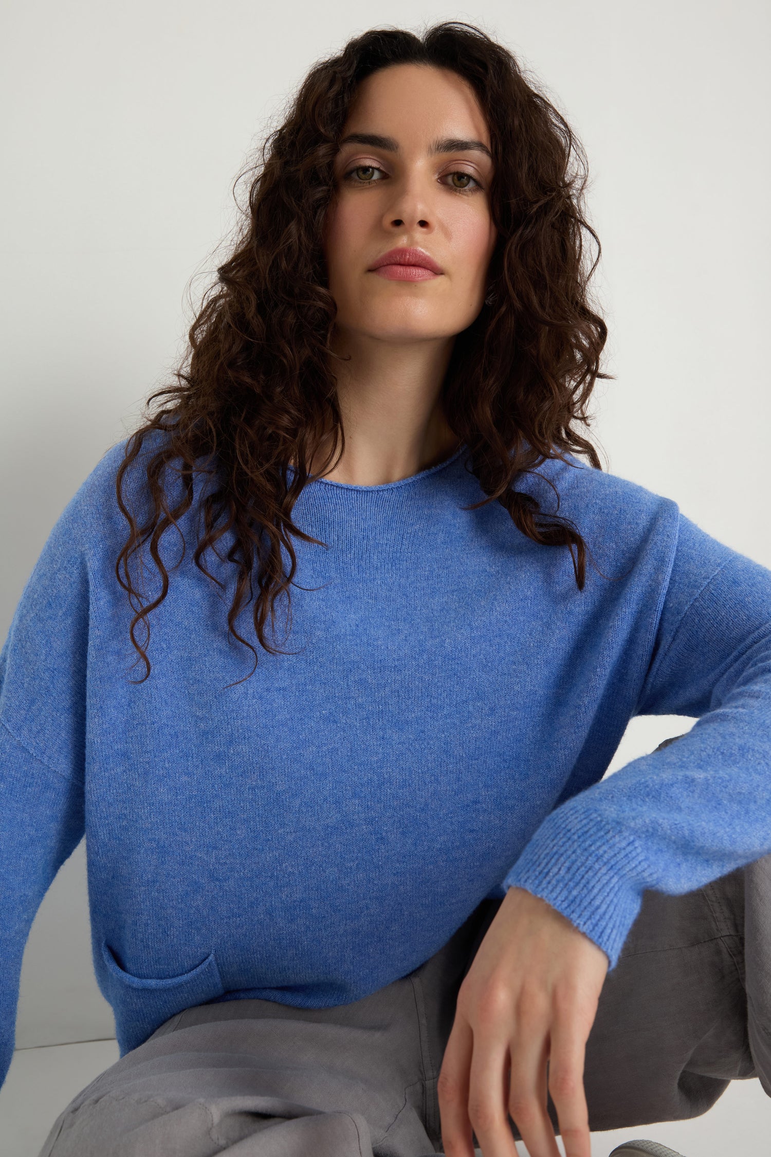 Wearing the Two Pocket Knit, a woman with long curly hair and gray pants sits against a plain white background—showcasing this blue sweater as an ideal piece for everyday layering.
