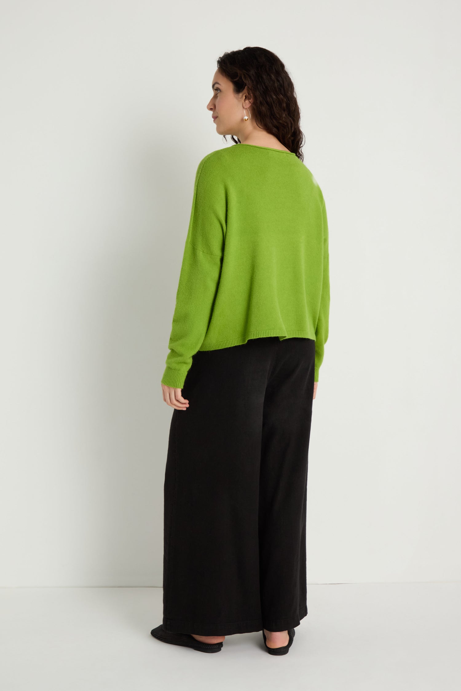 Woman stands against a plain white wall wearing the Two Pocket Knit—a relaxed women's sweater perfect for layering—paired with wide-leg black pants and black flats, facing away from the camera.