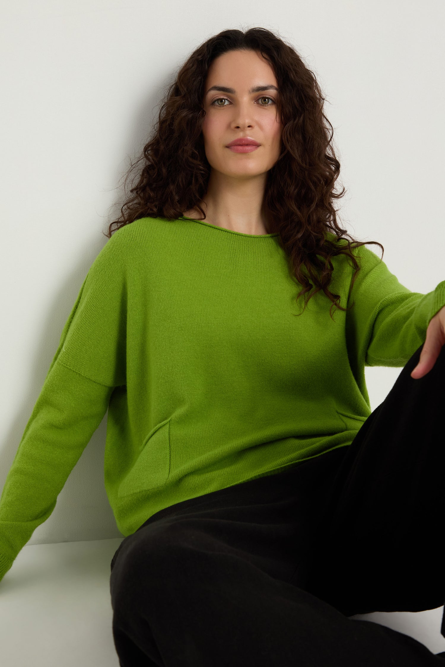 Woman with long curly hair wears a bright green Two Pocket Knit sweater and black pants, sitting against a white background—an ideal everyday layering piece.