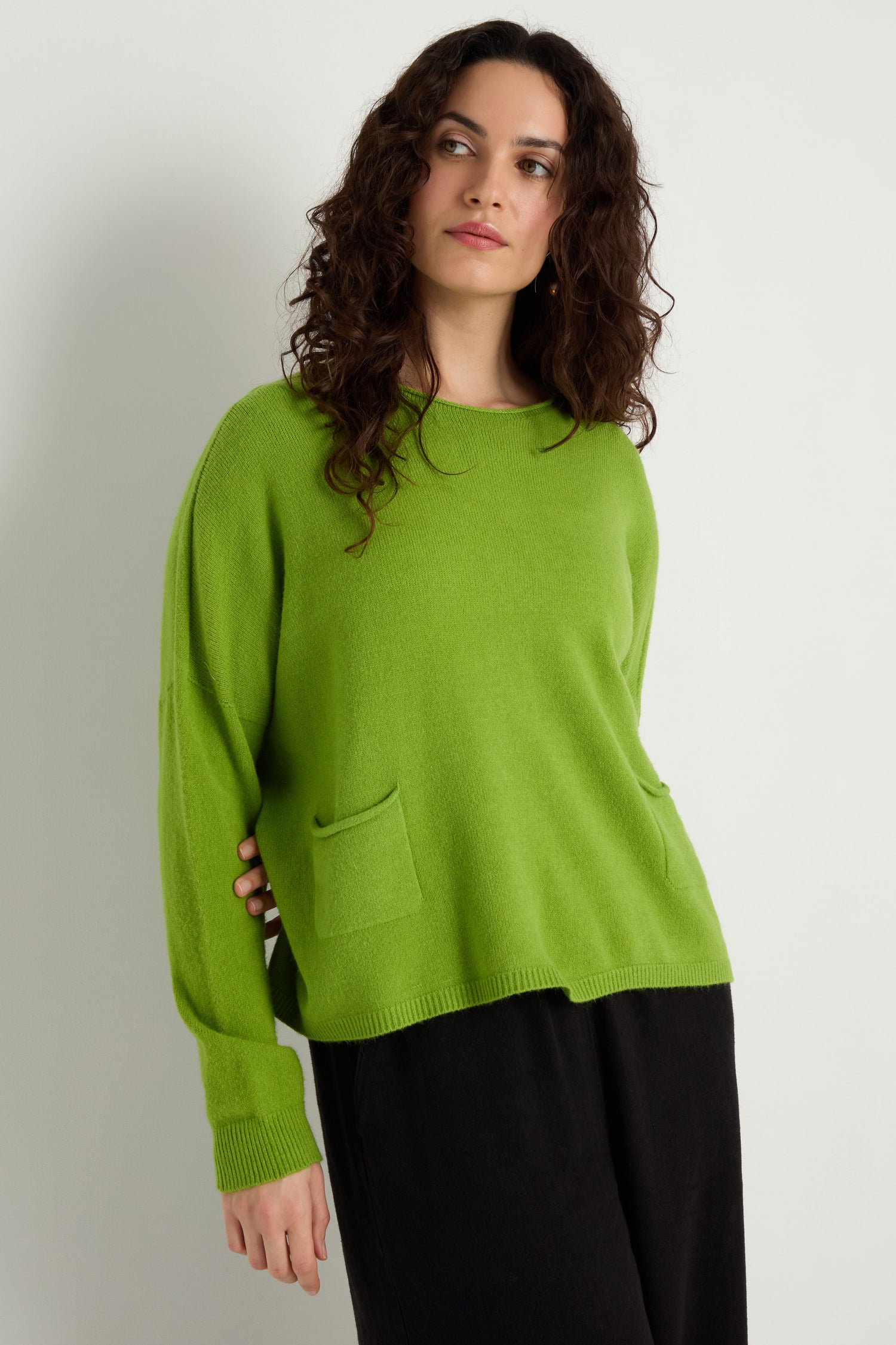 A person with long curly hair wears the Two Pocket Knit in bright green, featuring two front pockets, paired with black pants against a white background—an ideal layering piece for everyday wear.