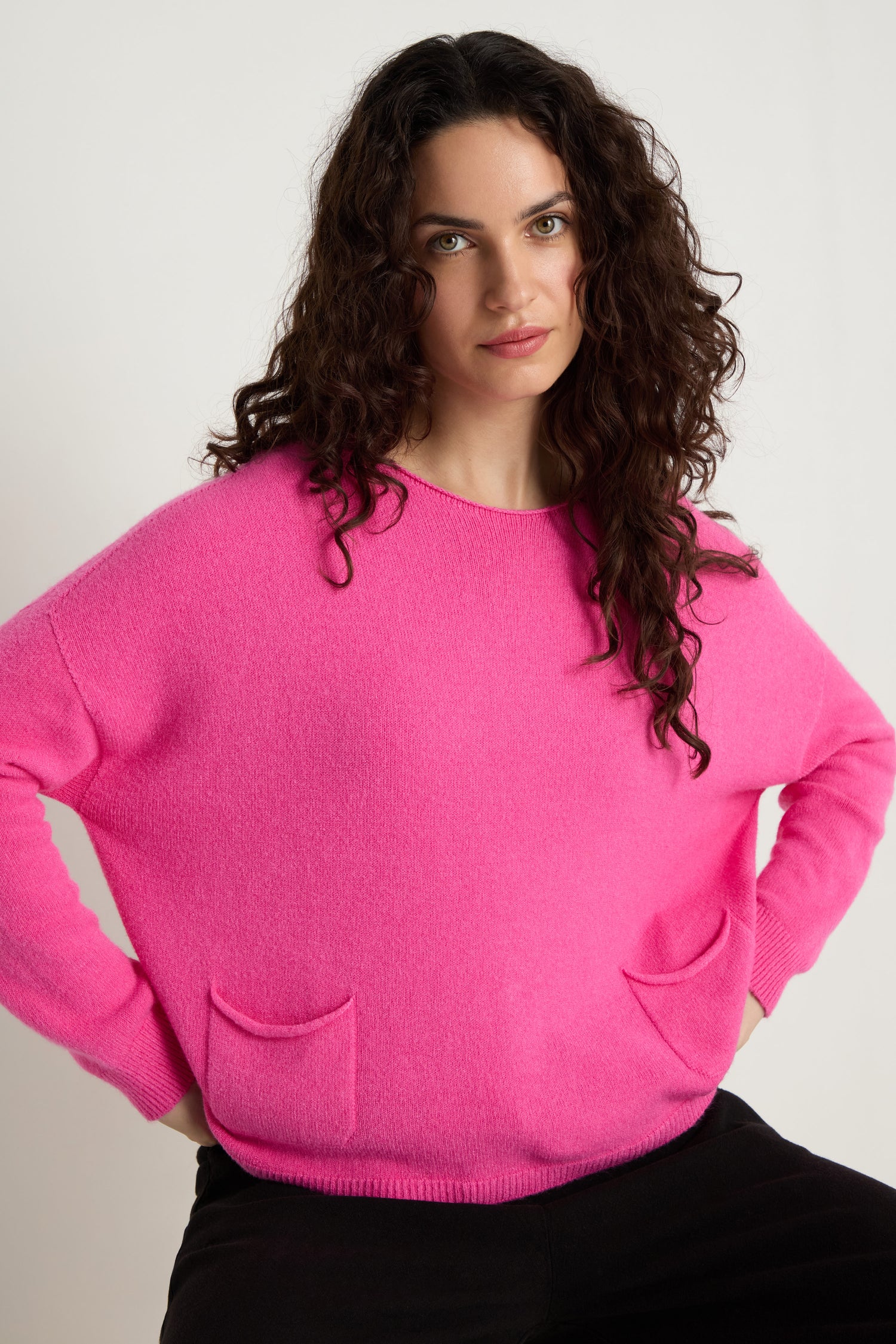 Woman with long curly hair wearing the Two Pocket Knit in bright pink, featuring two front pockets, posing with hands on hips against a plain background—an ideal layering piece for daily wear.