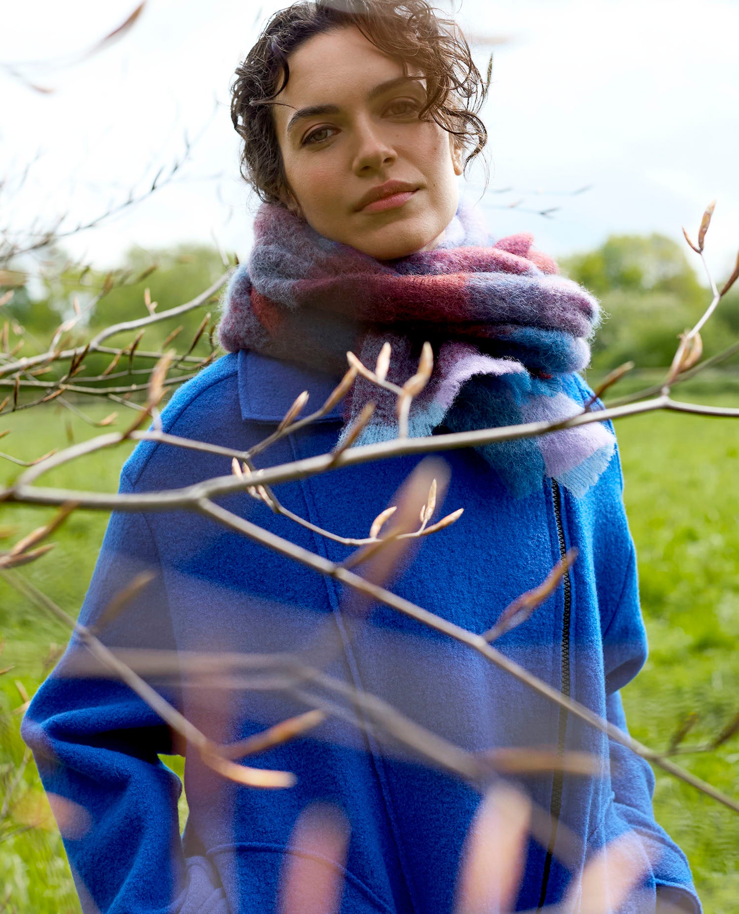 A woman in a blue coat and colourful scarf stands in a green field, framed by tree branches.