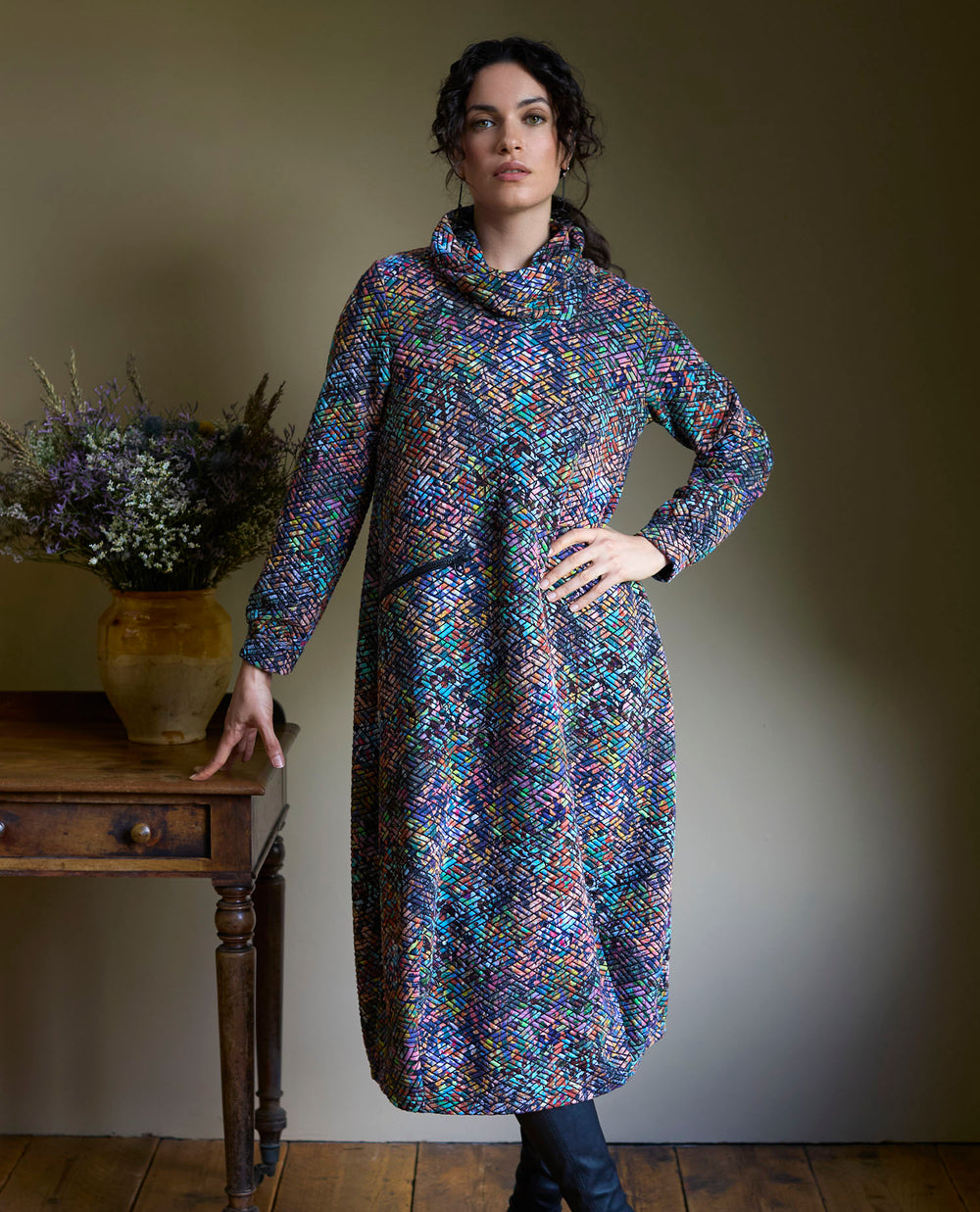 Woman in a colourful, patterned dress with a high collar, stands confidently in a room with wooden floors. A vase with flowers sits on a wooden table next to her.