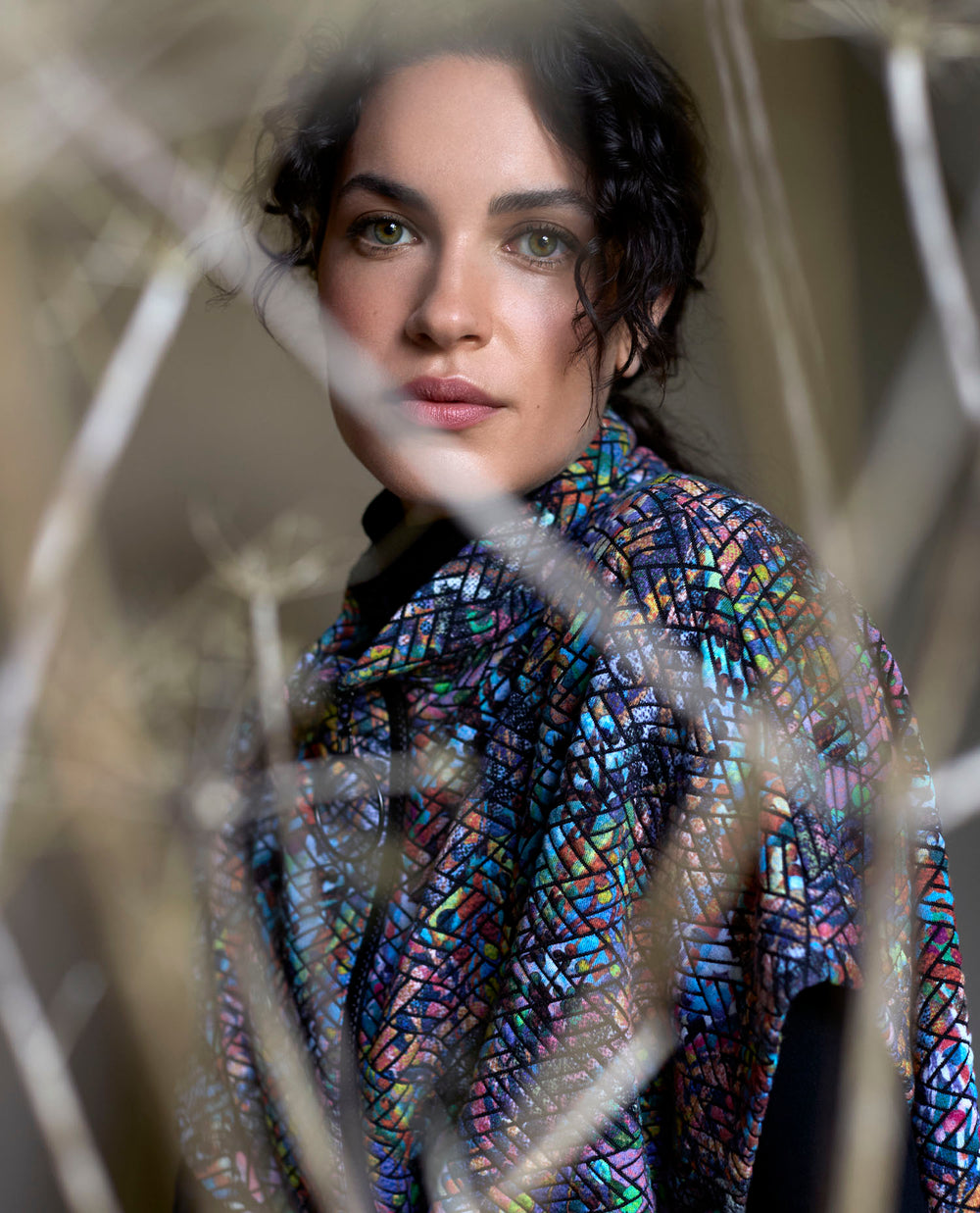 A woman with curly hair stands amid blurred branches, wearing a colourful, mosaic-patterned dress.
