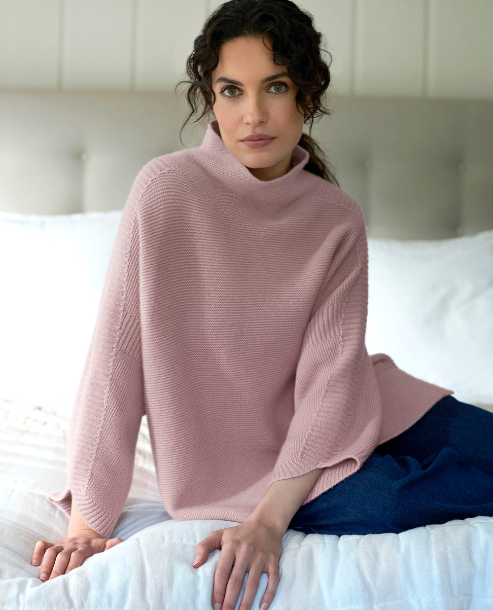 A woman with dark curly hair wearing a soft pink jumper and jeans sits on a bed with white sheets.