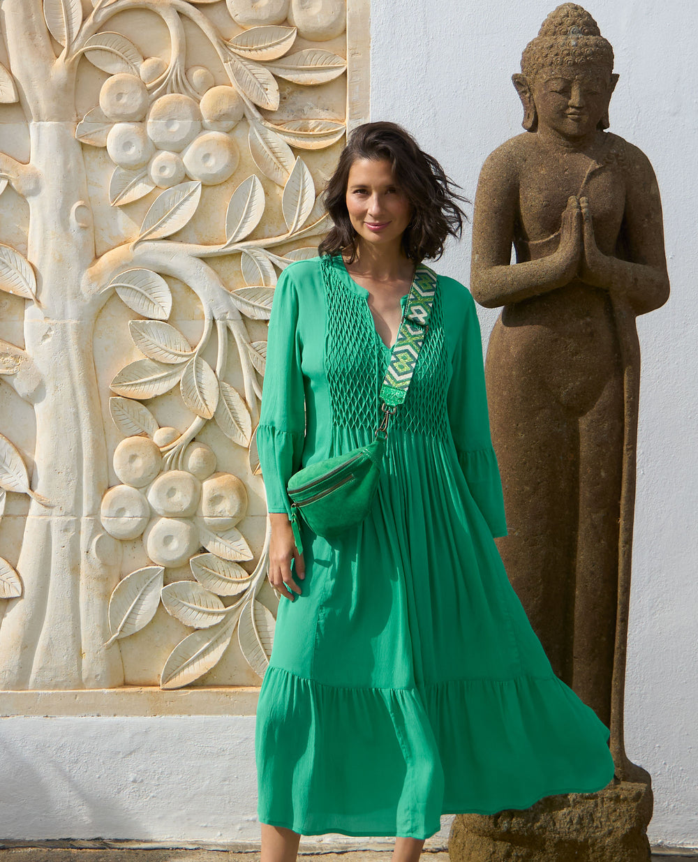 A woman in a flowing green dress stands beside a stone Buddha statue. A carved floral mural decorates the wall behind her.