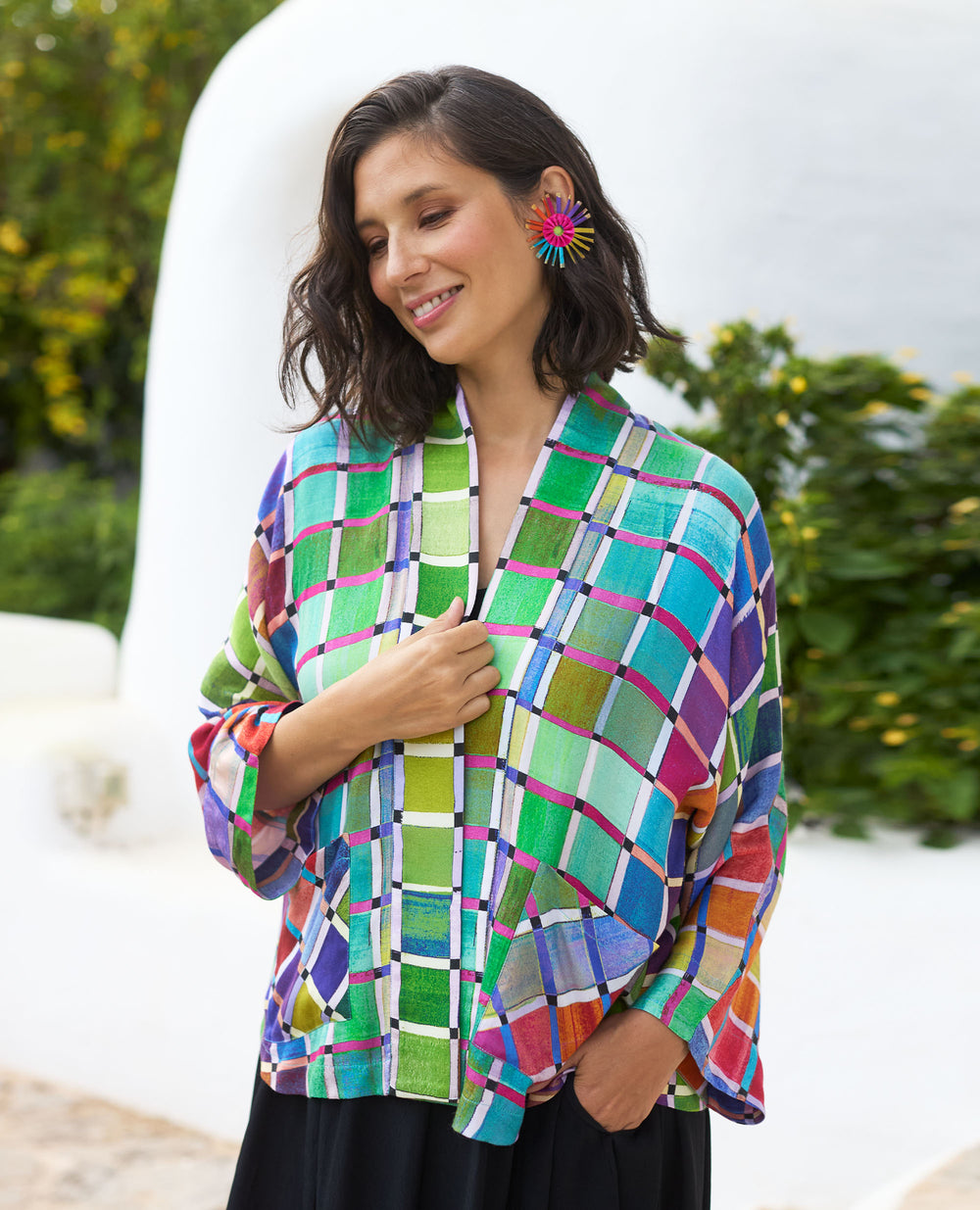 Woman smiling in a colourful, checkered jacket and vibrant earrings, standing outdoors against a bright backdrop with greenery.