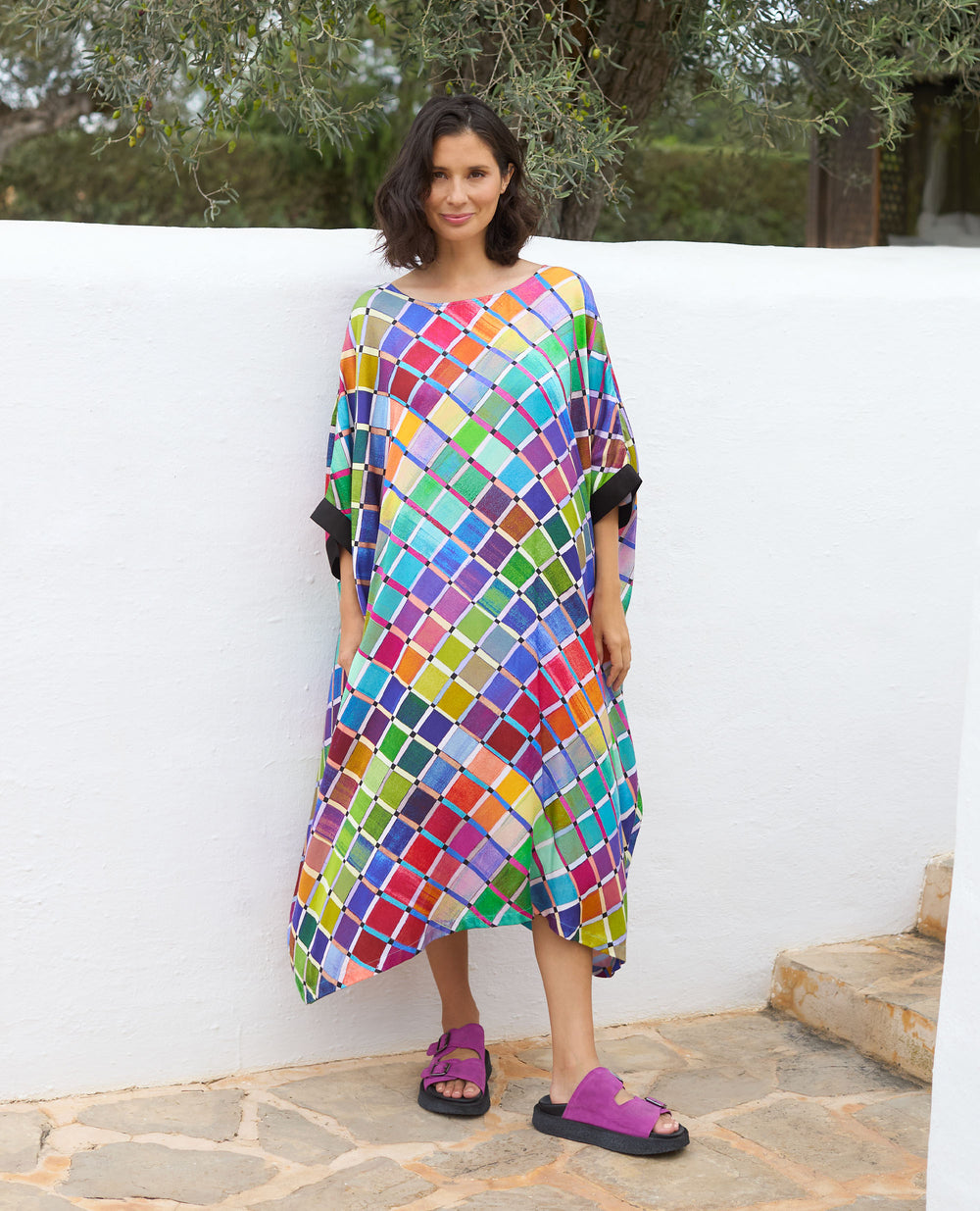 A woman stands against a white wall outside, she is wearing a colourful, geometric-patterned dress and purple sandals.