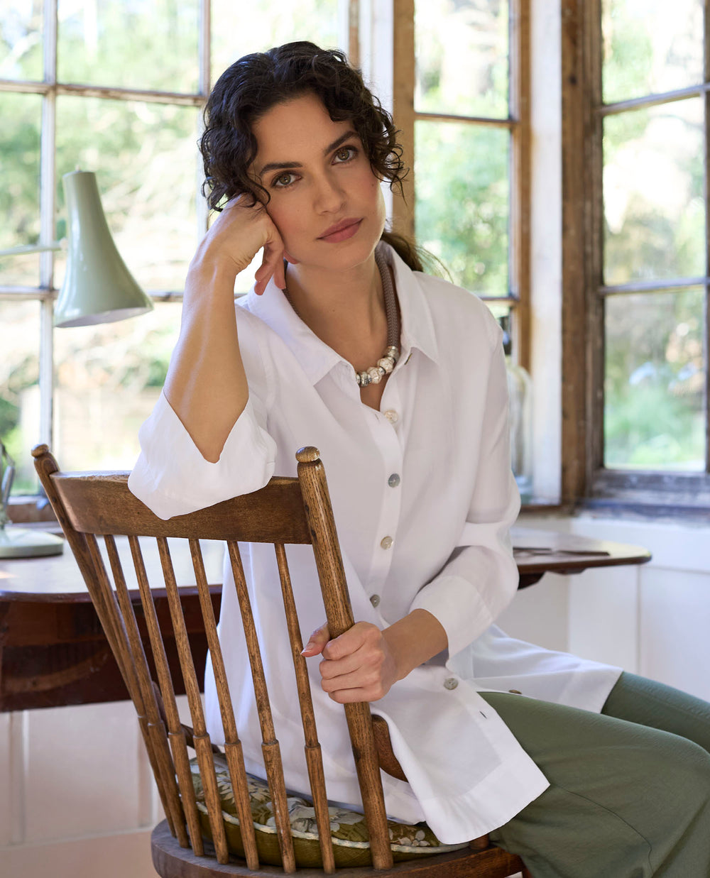 A woman with curly hair sits pensively on a wooden chair by a bright window, she is wearing a white blouse and green trousers.
