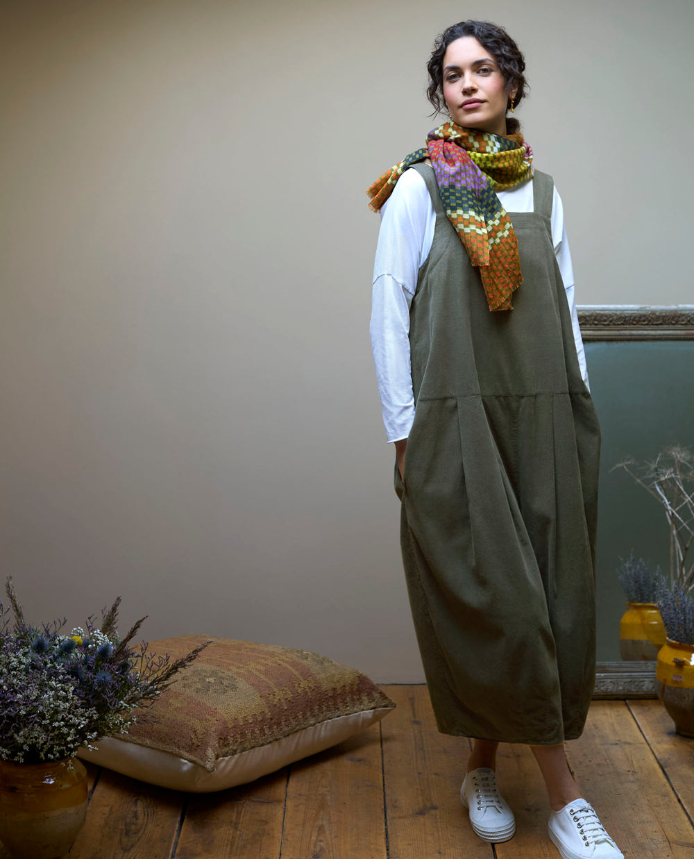 A woman in an olive green dress and colourful scarf stands on a wooden floor, next to a cushion and dried plants.
