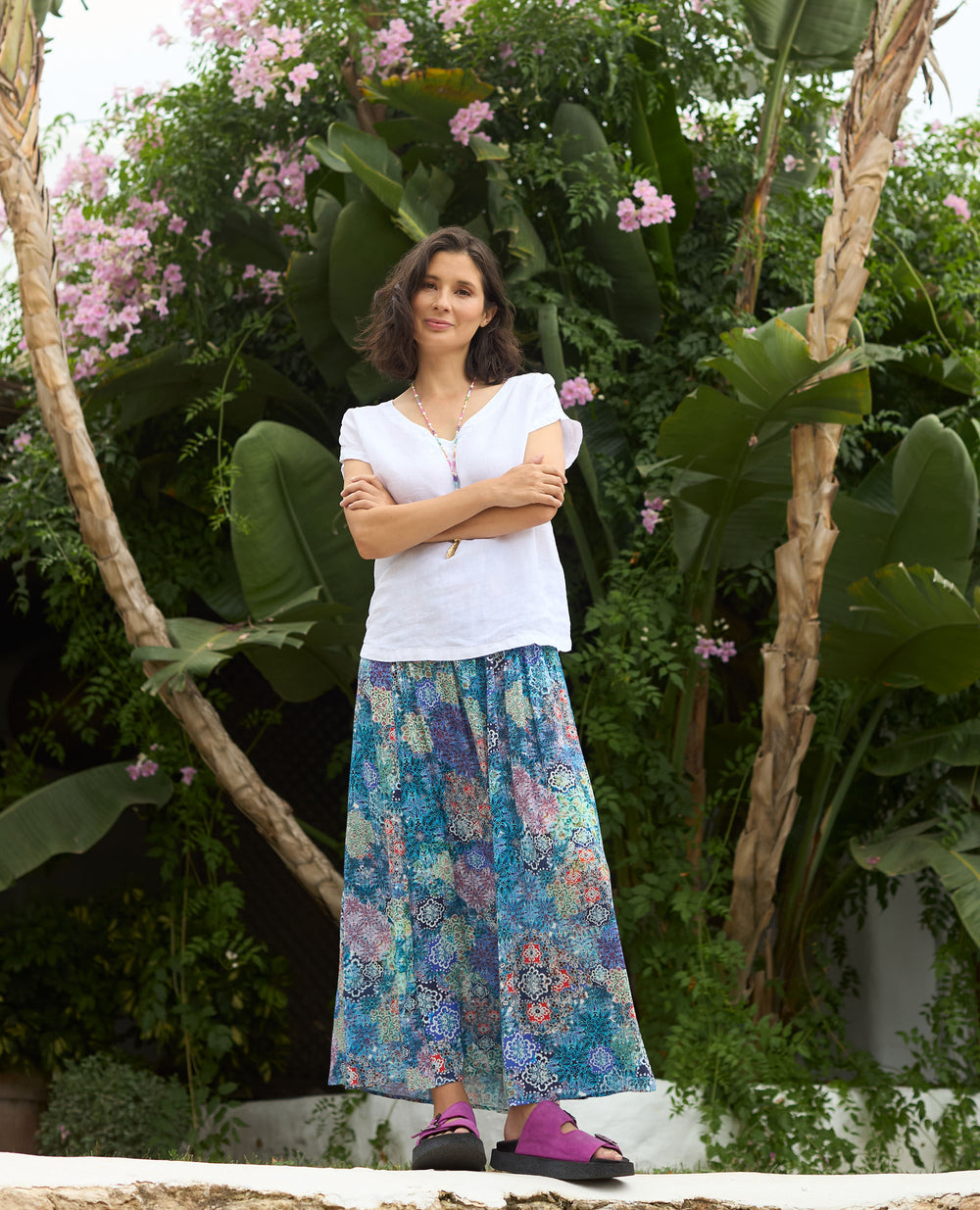 A woman stands calmly in front of green foliage with pink flowers. She's wearing a white top, colourful floral skirt, and purple sandals.