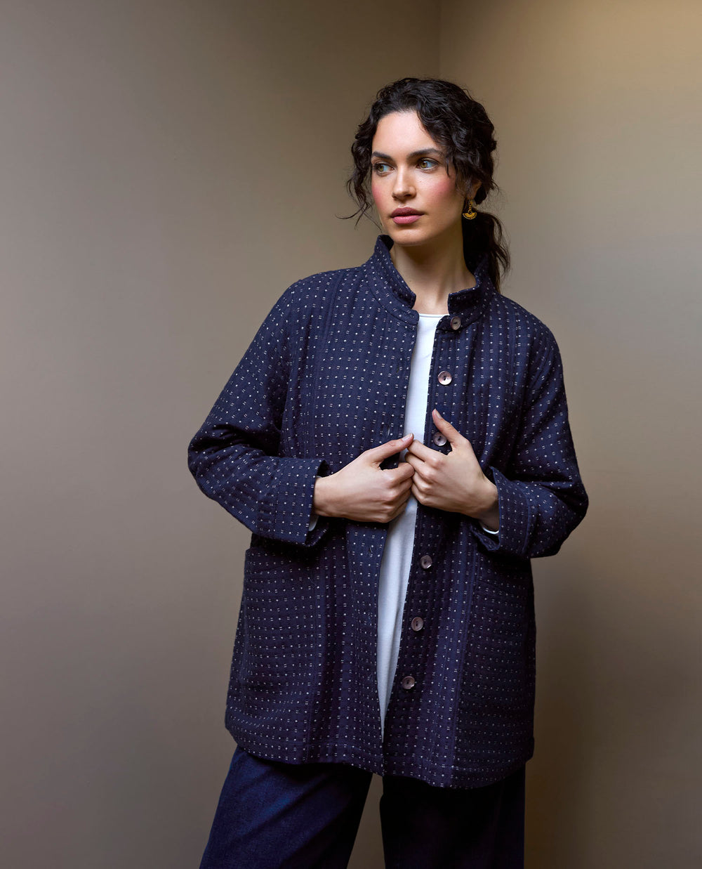 A woman with wavy hair in a navy, polka-dotted jacket and white top stands against a neutral background.