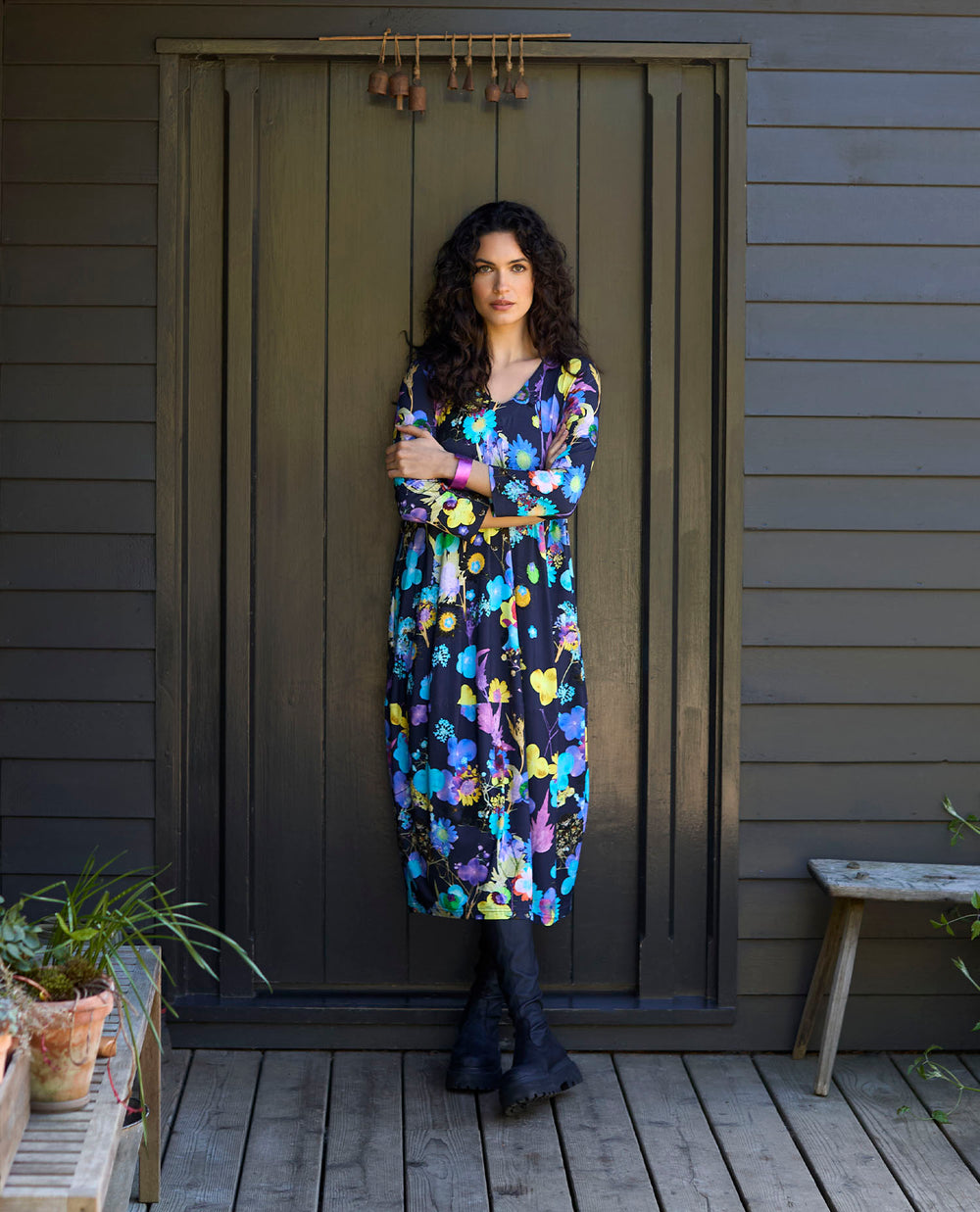 A woman stands confidently against a dark wooden door, wearing a colourful floral dress and black boots.