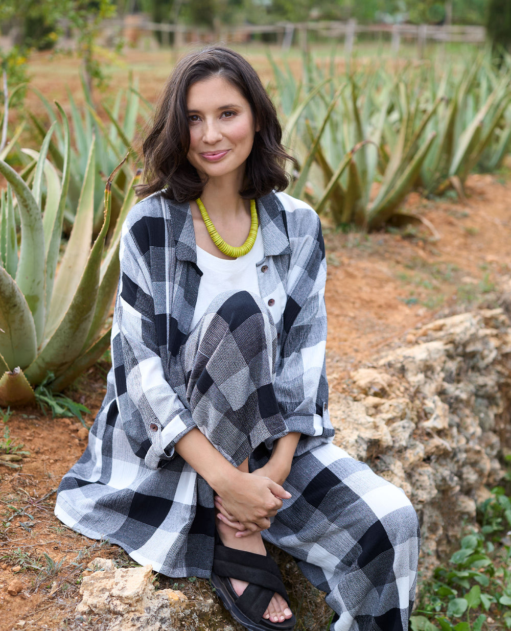 A woman in a black-and-white checkered outfit sits on a stone ledge, surrounded by aloe plants.