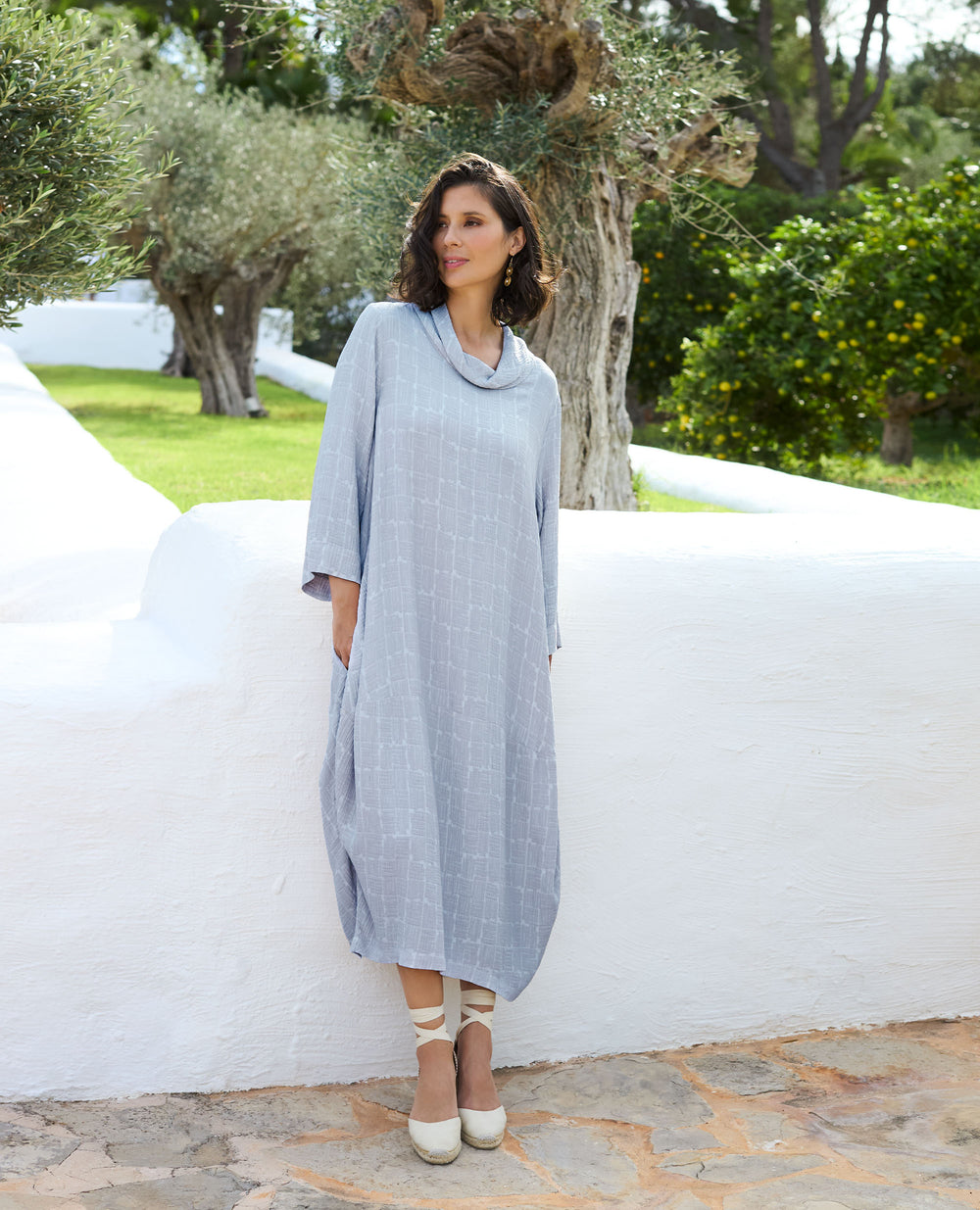 A woman in a light grey, loose dress stands against a white wall outdoors. She is on a stone path, with green trees in the background.