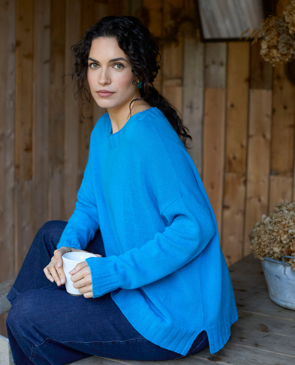 A woman with wavy dark hair in a bright blue jumper sits on a wooden bench, holding a white mug. The background features rustic wooden walls and dried flowers.