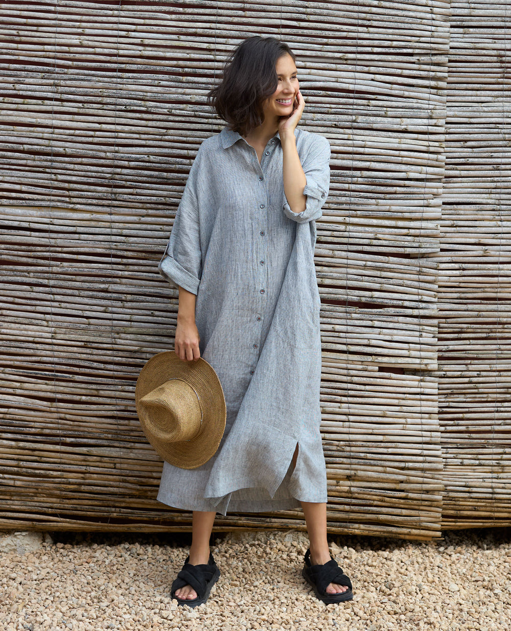 A woman in a relaxed stance, wearing a button-down grey dress and sandals, holds a straw hat by her side. She's smiling, standing against a rustic wooden fence.