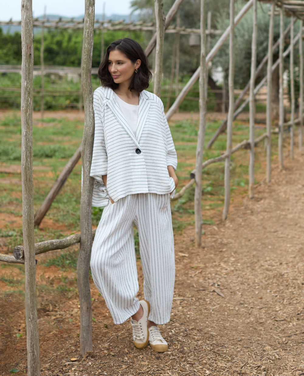 A woman in a loose, white pinstripe suit leans casually against a wooden structure in a garden, surrounded by greenery.
