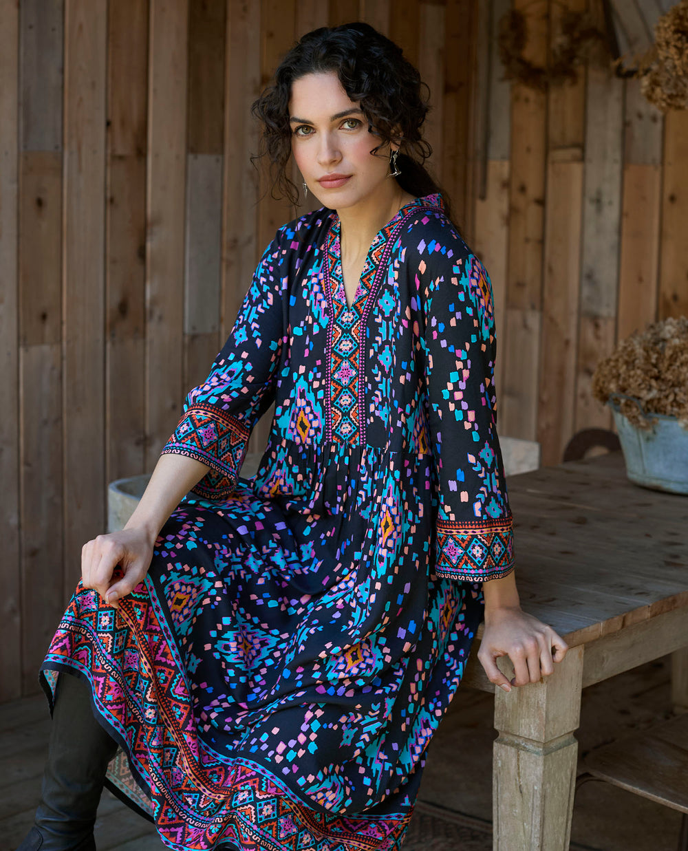 A woman in a patterned dress sits on a rustic wooden table in a wooden outdoor area. The dress features vibrant geometric designs.