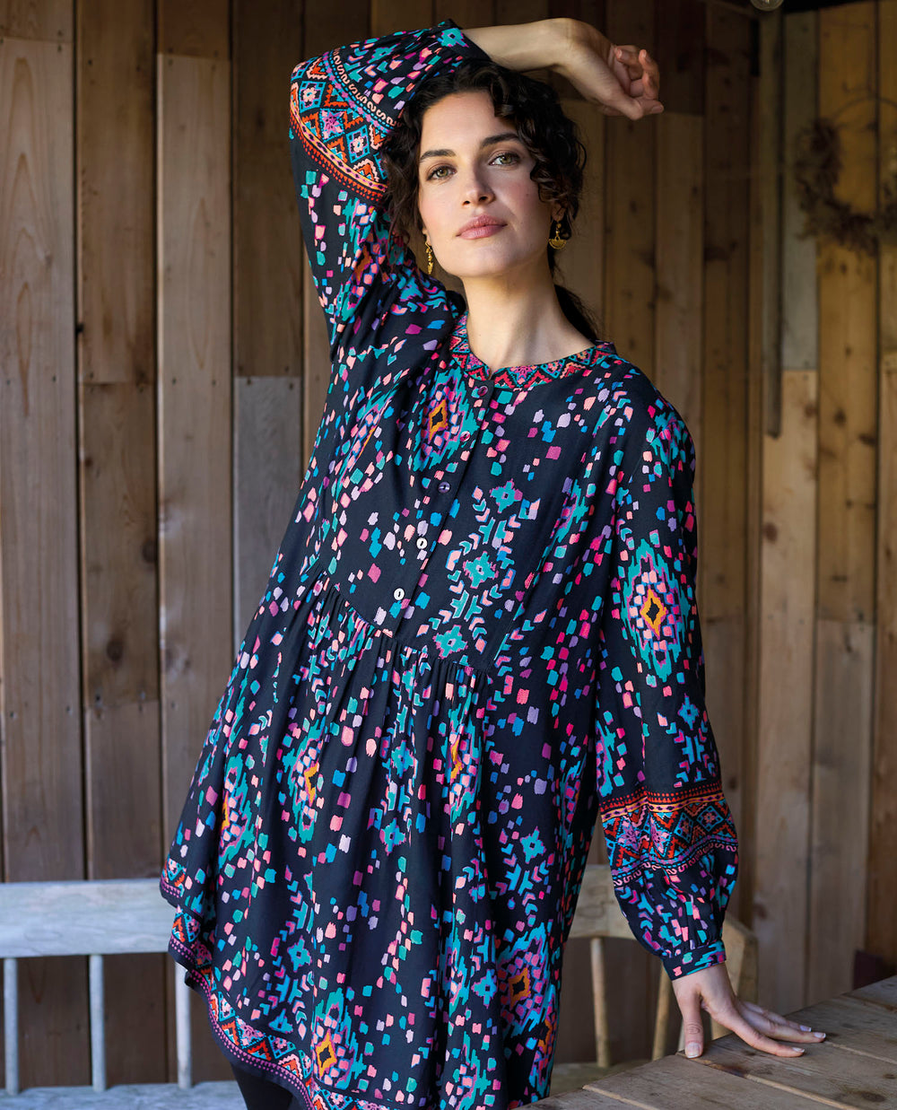 A woman in a patterned dress sits on a rustic wooden table in a wooden outdoor area posing with her hand over her head. The dress features vibrant geometric designs.
