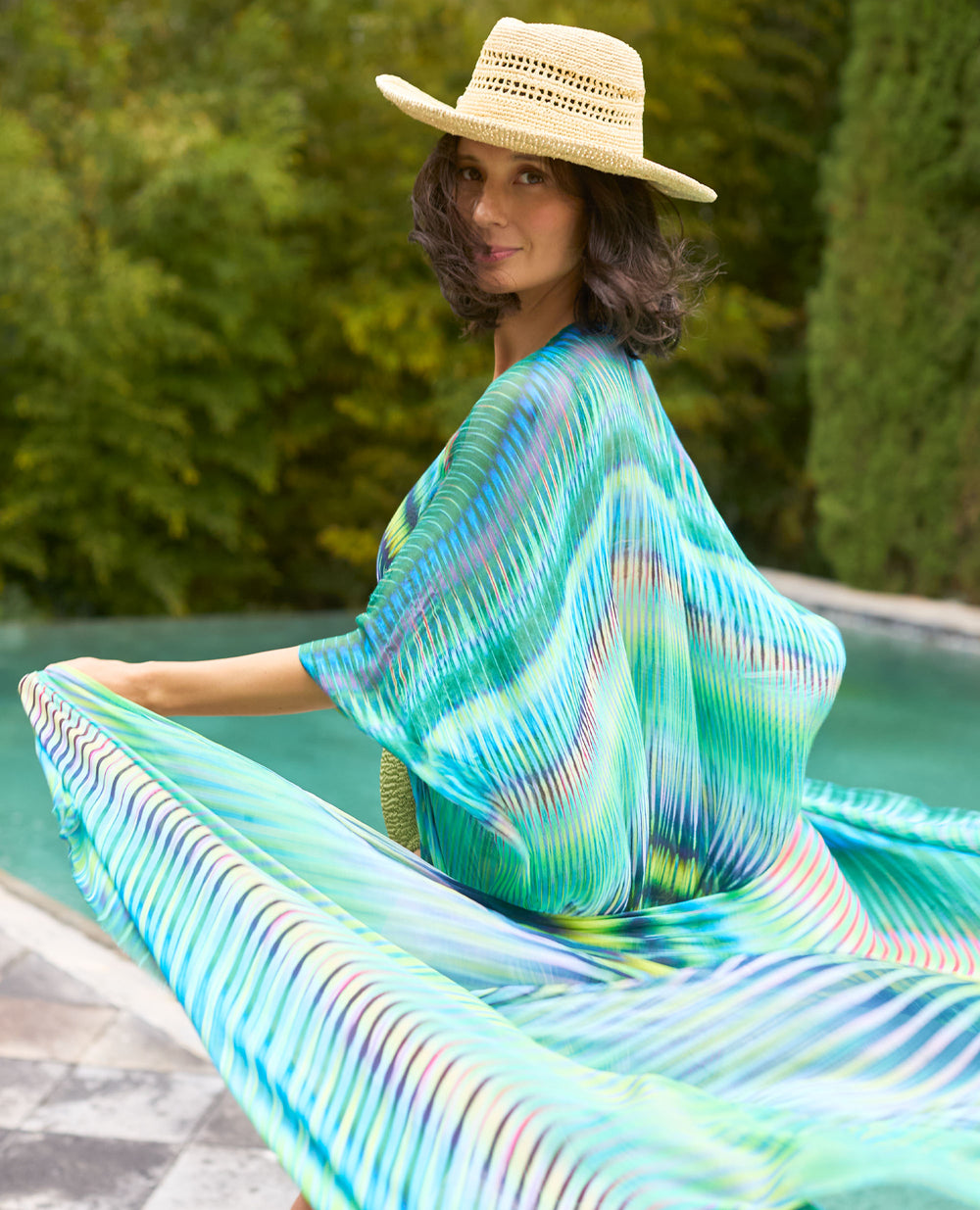 A woman in a straw hat and colourful, flowing dress smiles by a pool with greenery in the background.