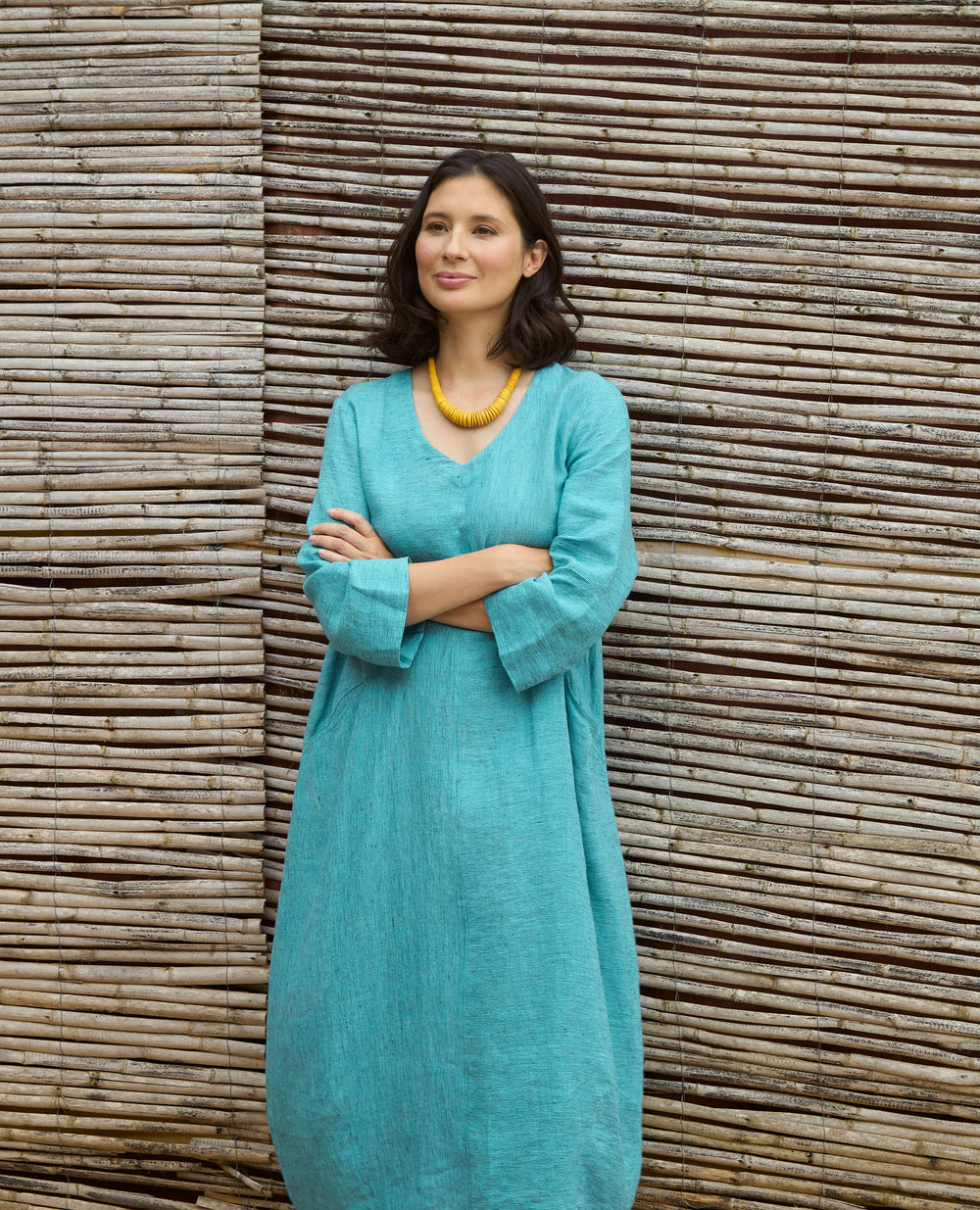 A woman in a teal dress with yellow necklace, stands confidently with arms crossed in front of a rustic bamboo wall.
