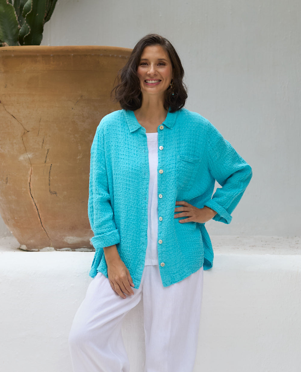 A smiling woman in a turquoise button-up shirt and white trousers stands beside a large terracotta pot in a bright outdoor area.