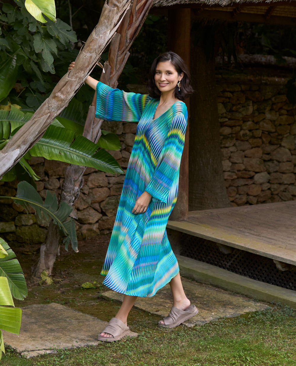 A smiling woman in a turquoise button-up shirt and white trousers stands beside a large terracotta pot in a bright outdoor area.