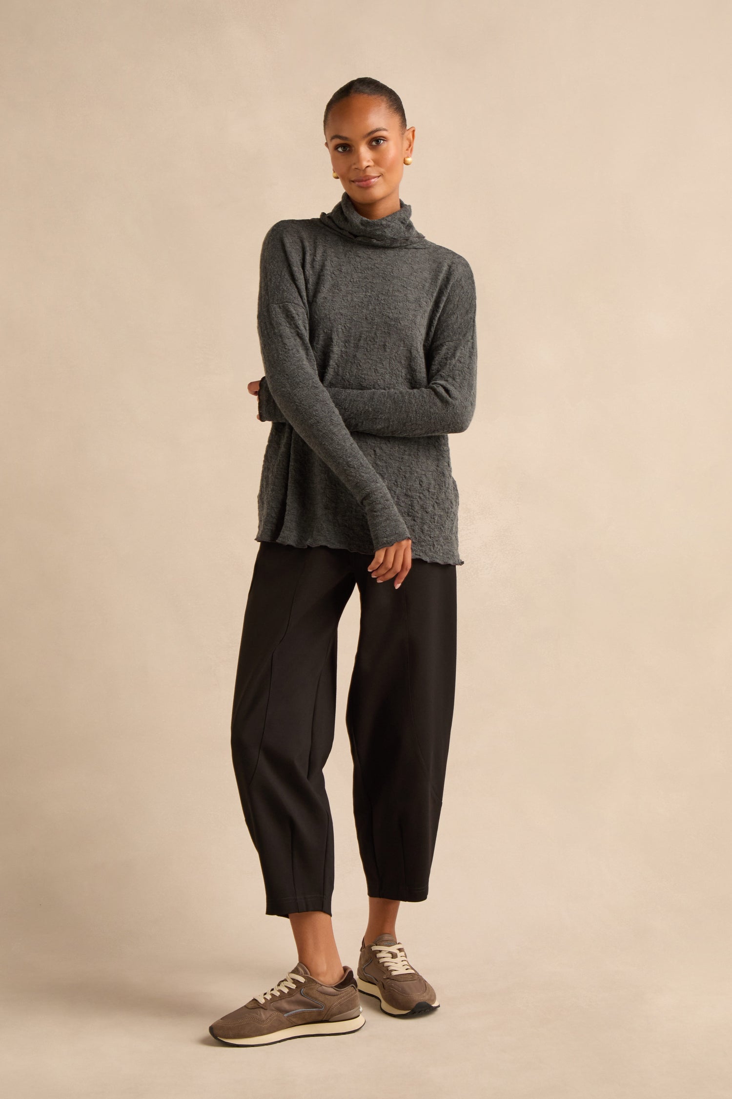 A woman stands against a beige background, wearing the Anthracite Textured Merino Roll Neck Top—crafted in Denmark—paired with black cropped pants and brown sneakers, her arms relaxed and one foot slightly forward.