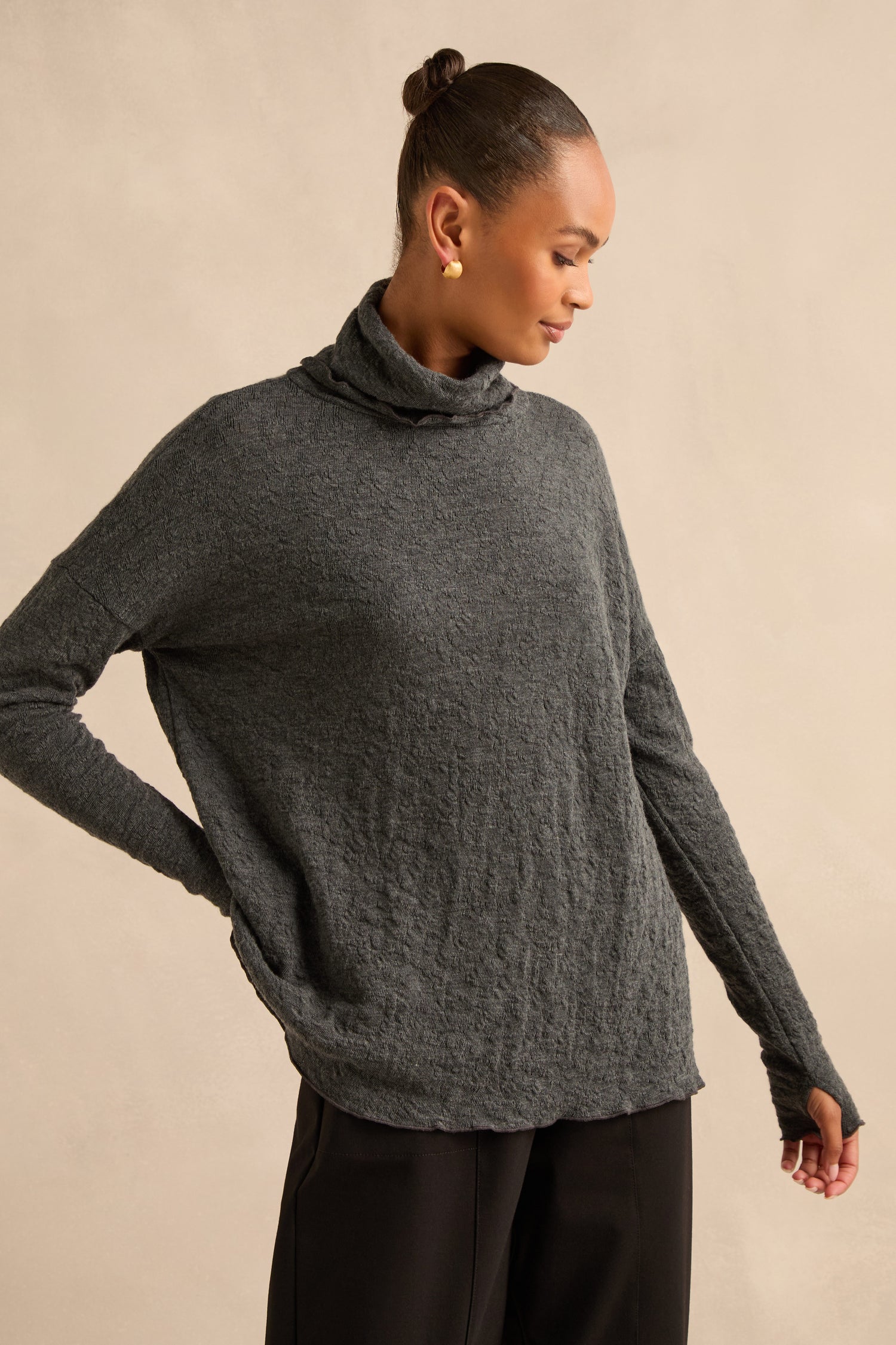 A woman stands against a neutral background, looking to the side with her hair in a bun and one hand on her hip, wearing the Anthracite Textured Merino Roll Neck Top paired with black pants.